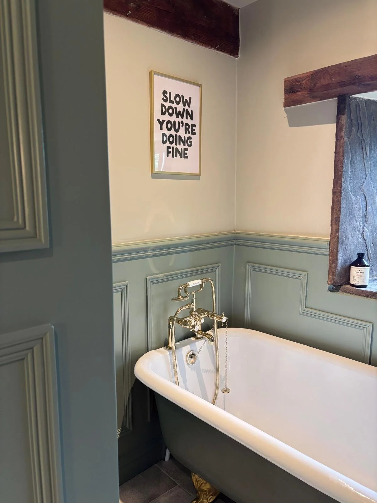 A bathroom with a black clawfoot tub, olive green wainscoting, a framed sign that says 'Slow down you're doing fine', a small shelf with a soap bottle, and a window with a stone frame.