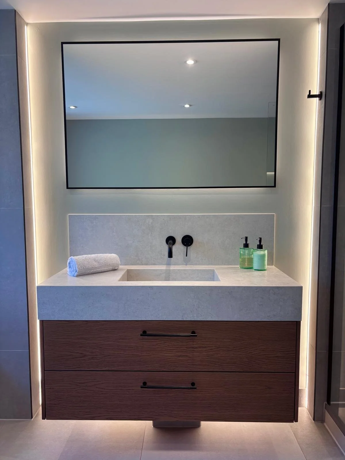 Modern bathroom vanity with a large rectangular mirror, a round white towel, and two green soap dispensers, with LED lighting around the mirror.