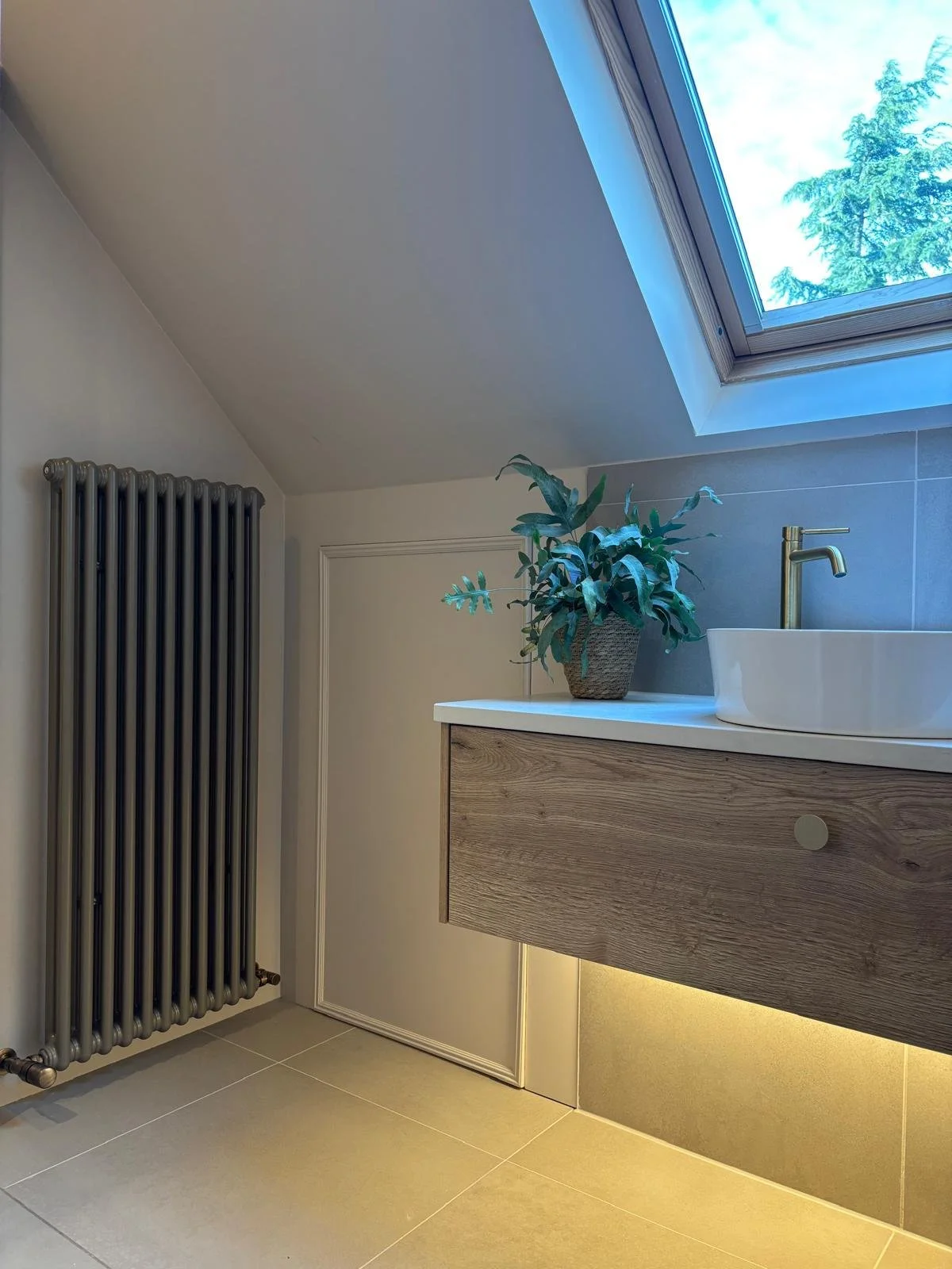 Modern bathroom with sloped ceiling, skylight window showing blue sky and trees, a sleek radiator on the wall, a potted plant on a wooden vanity, white vessel sink, and brass faucet.