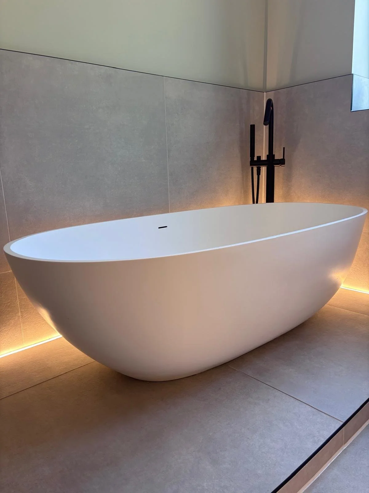 Free-standing white bathtub with a black floor-mounted faucet in a modern bathroom with beige tiles and minimal lighting.