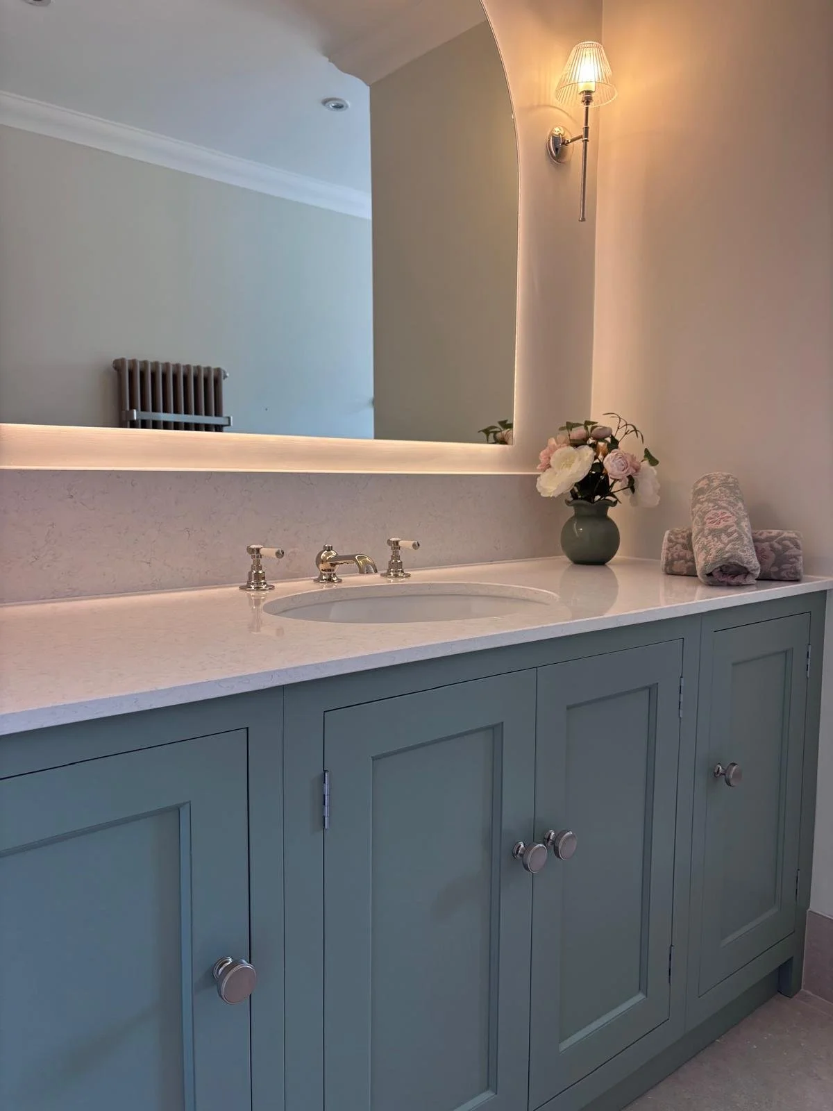 Bathroom vanity with a large mirror, a green vase with pink and white flowers, and rolled towels, with soft lighting and cabinets below.