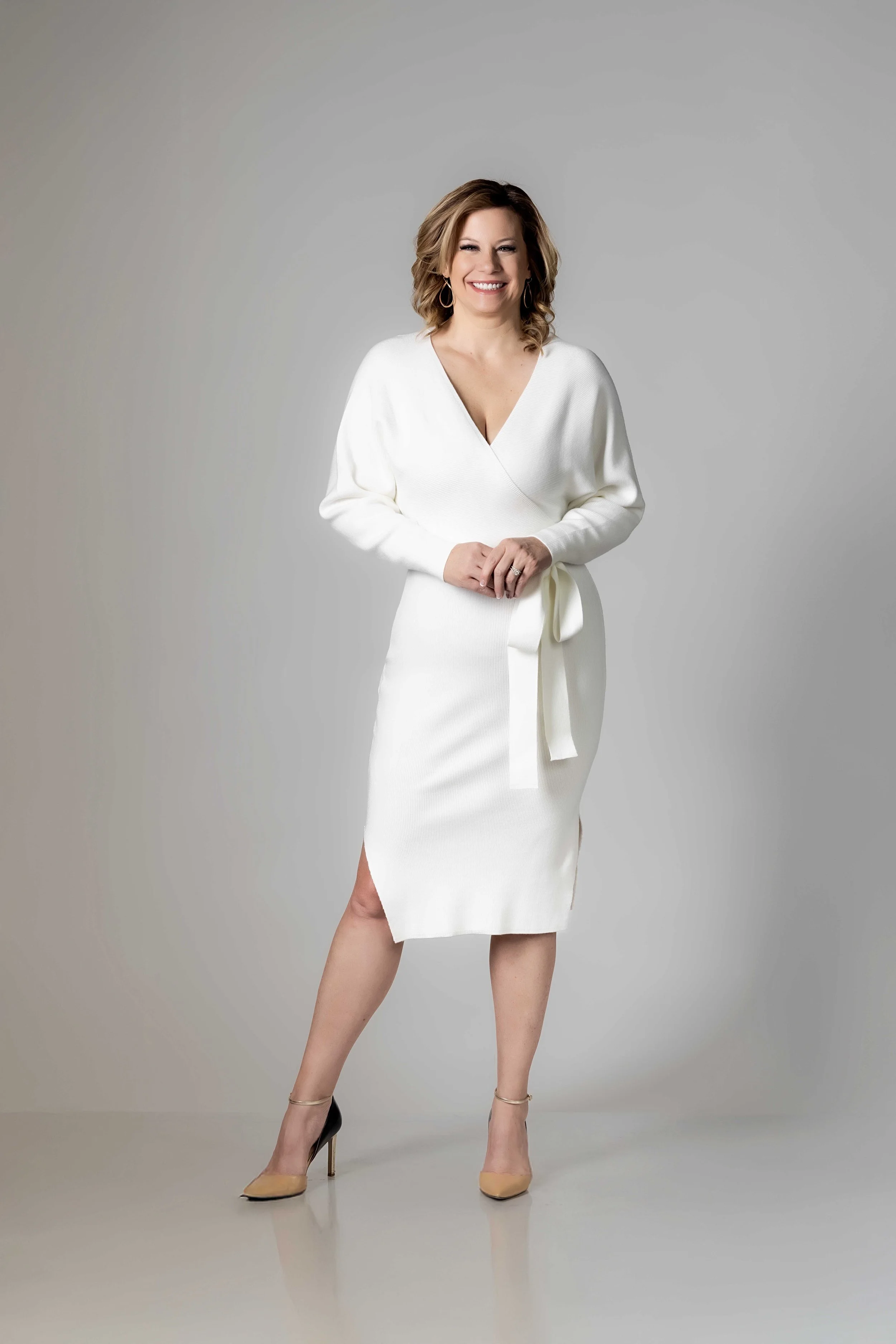 A woman with shoulder-length brown hair, blue eyes, and a bright smile, wearing a black textured dress with her left hand in her pocket standing indoors near white flowers.