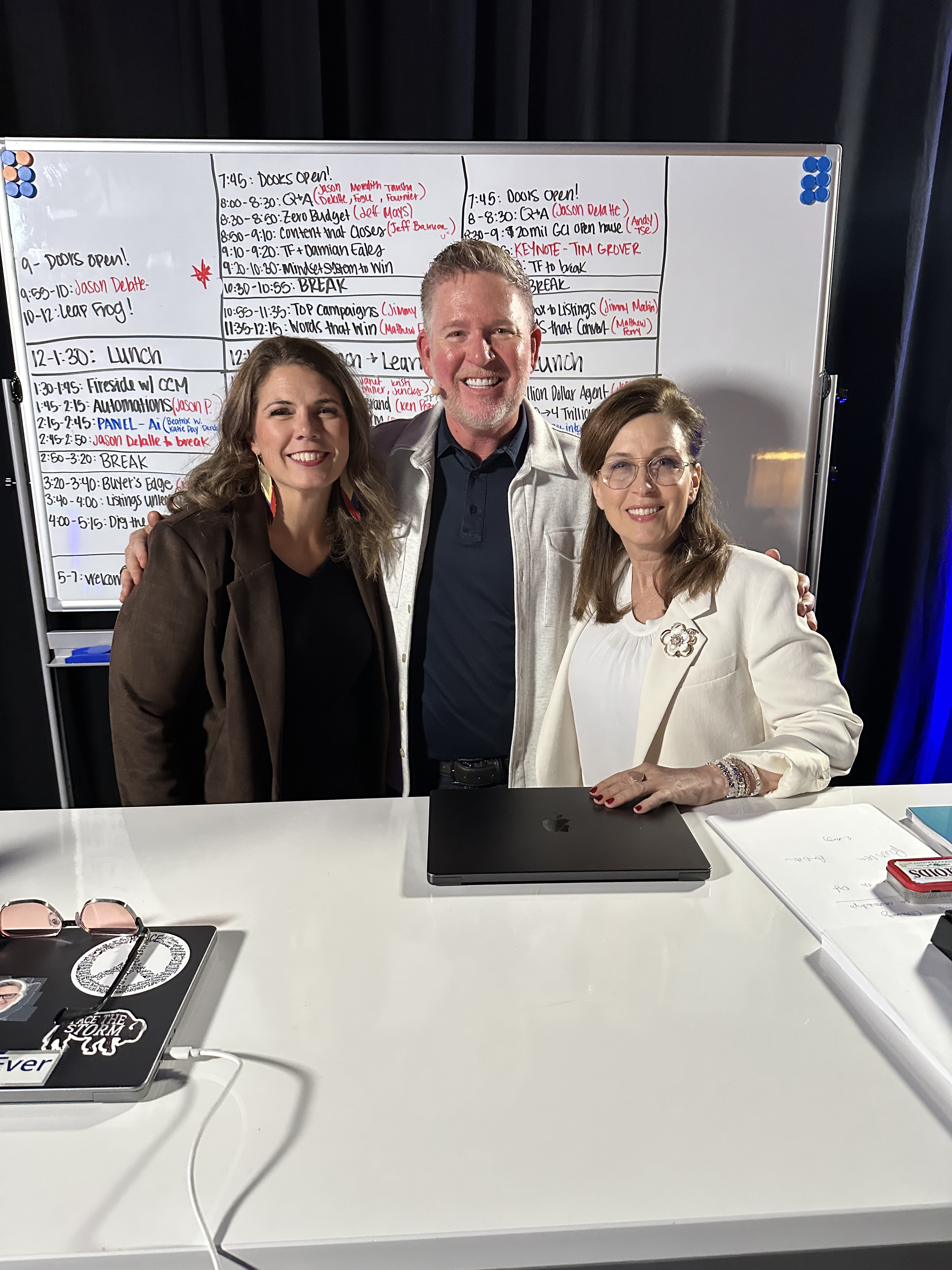 Three people standing behind a white table with laptops and documents, smiling, with a large whiteboard filled with handwritten schedules and notes behind them.
