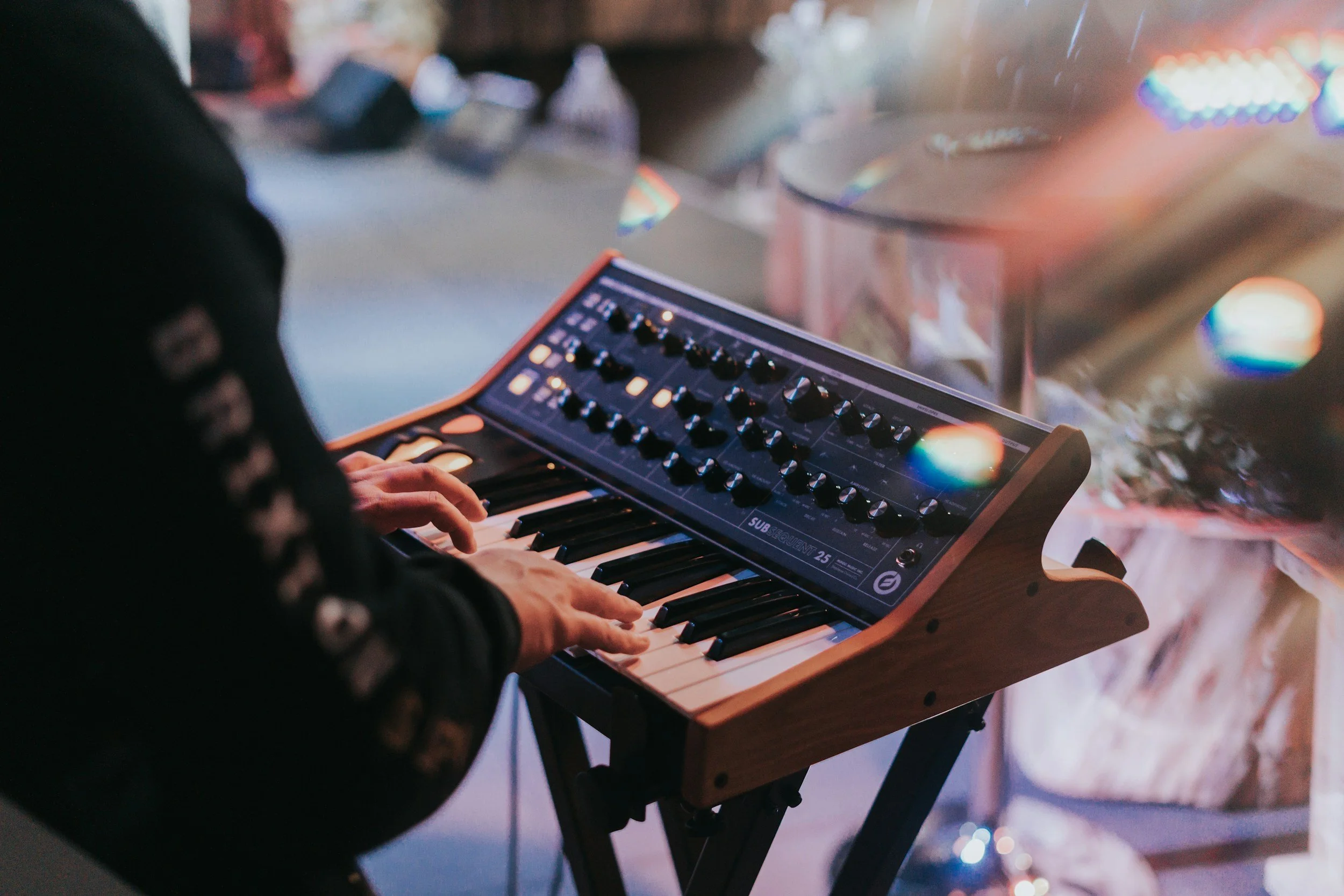 Nahaufnahme eines digitalen Synthesizers mit Regler und Tasten, gespielt von einer Person in einer Raum mit bunter Beleuchtung.