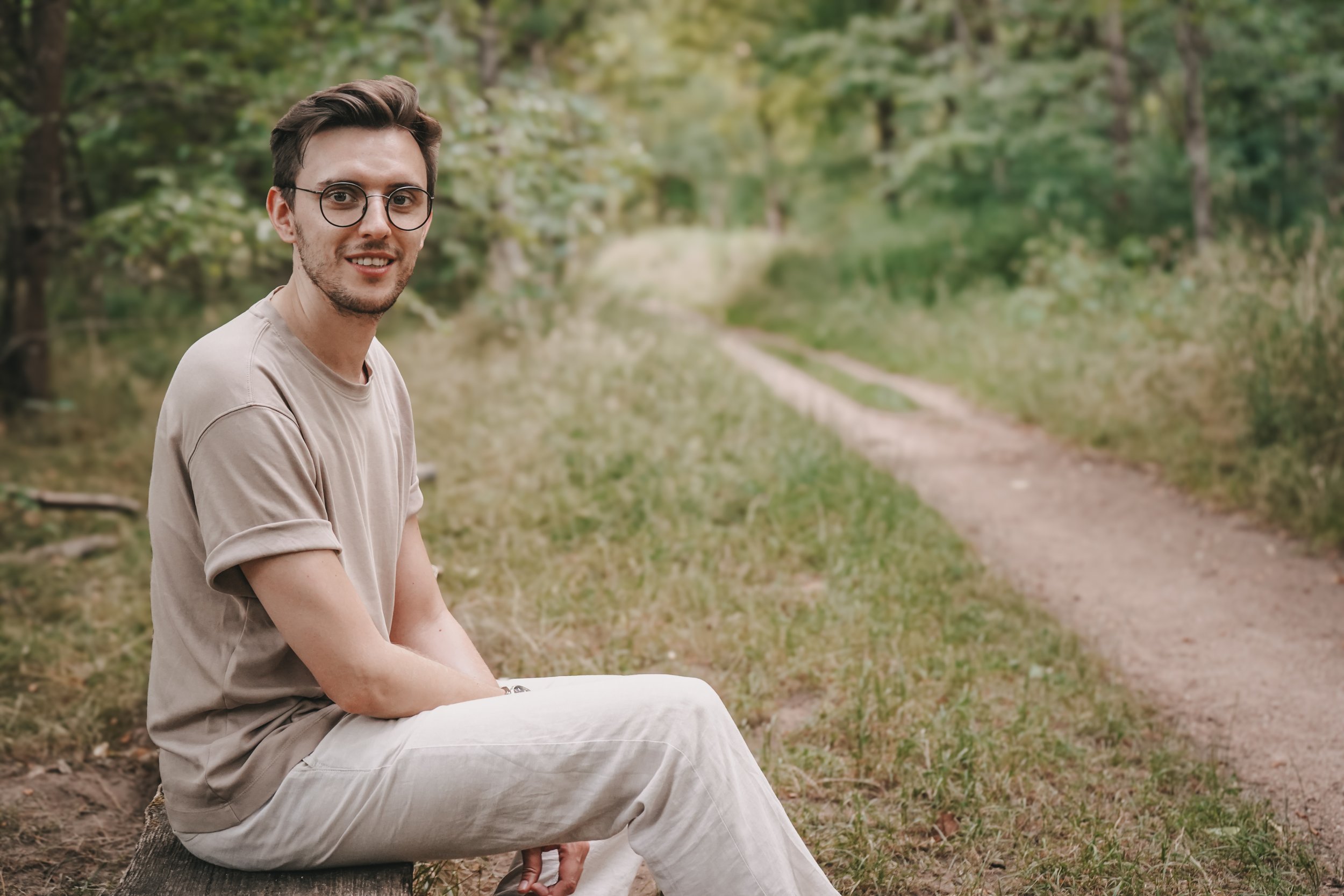 Ein junger Mann mit dunklen Haaren, Brille und legerer Kleidung sitzt auf einem Baumstumpf im Grünen, Blick Richtung Kamera.
