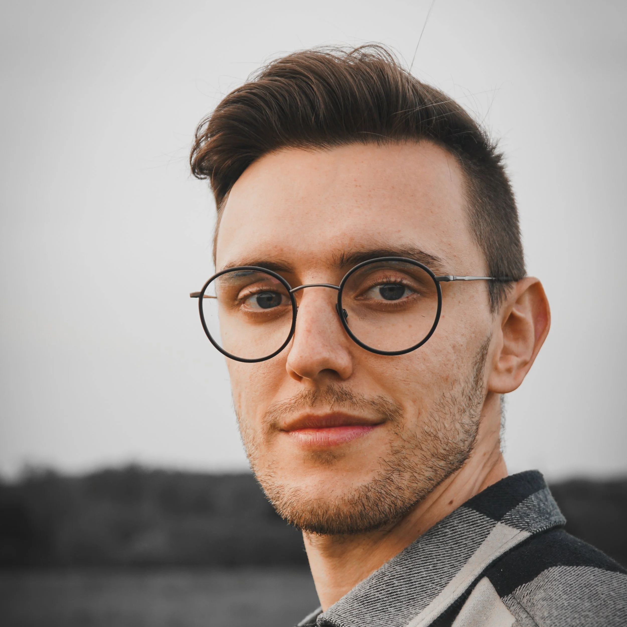 Porträt eines jungen Mannes mit Brille, dunklem Haar und Bart, in einem grauen Hemd, draußen mit unscharfem Hintergrund.
