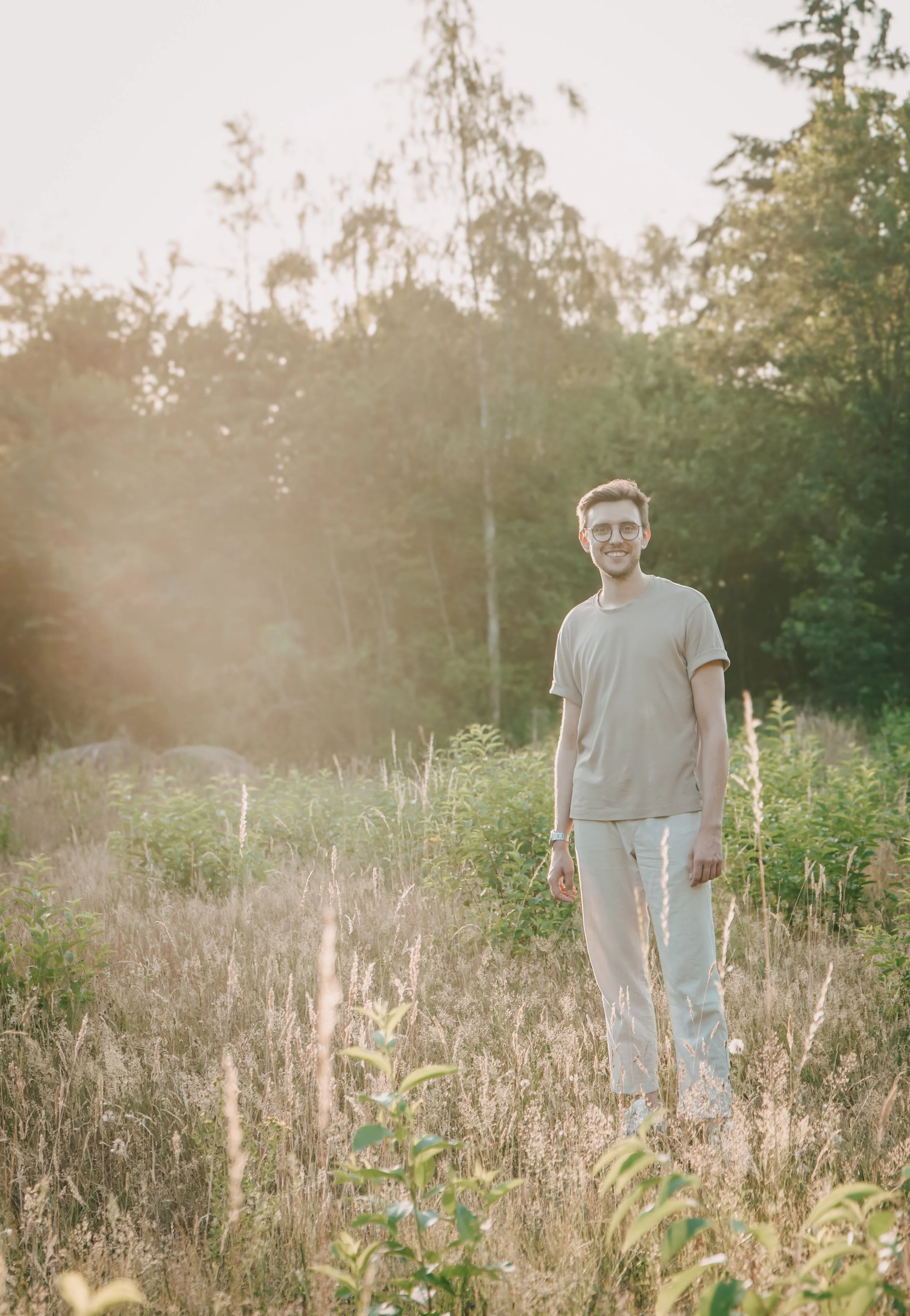 Ein lächelnder junger Mann mit Brille trägt ein helles T-Shirt und weiße Hosen, steht in einem offenen Feld mit hohen Gräsern und Büschen, umgeben von Bäumen, bei Sonnenuntergang.