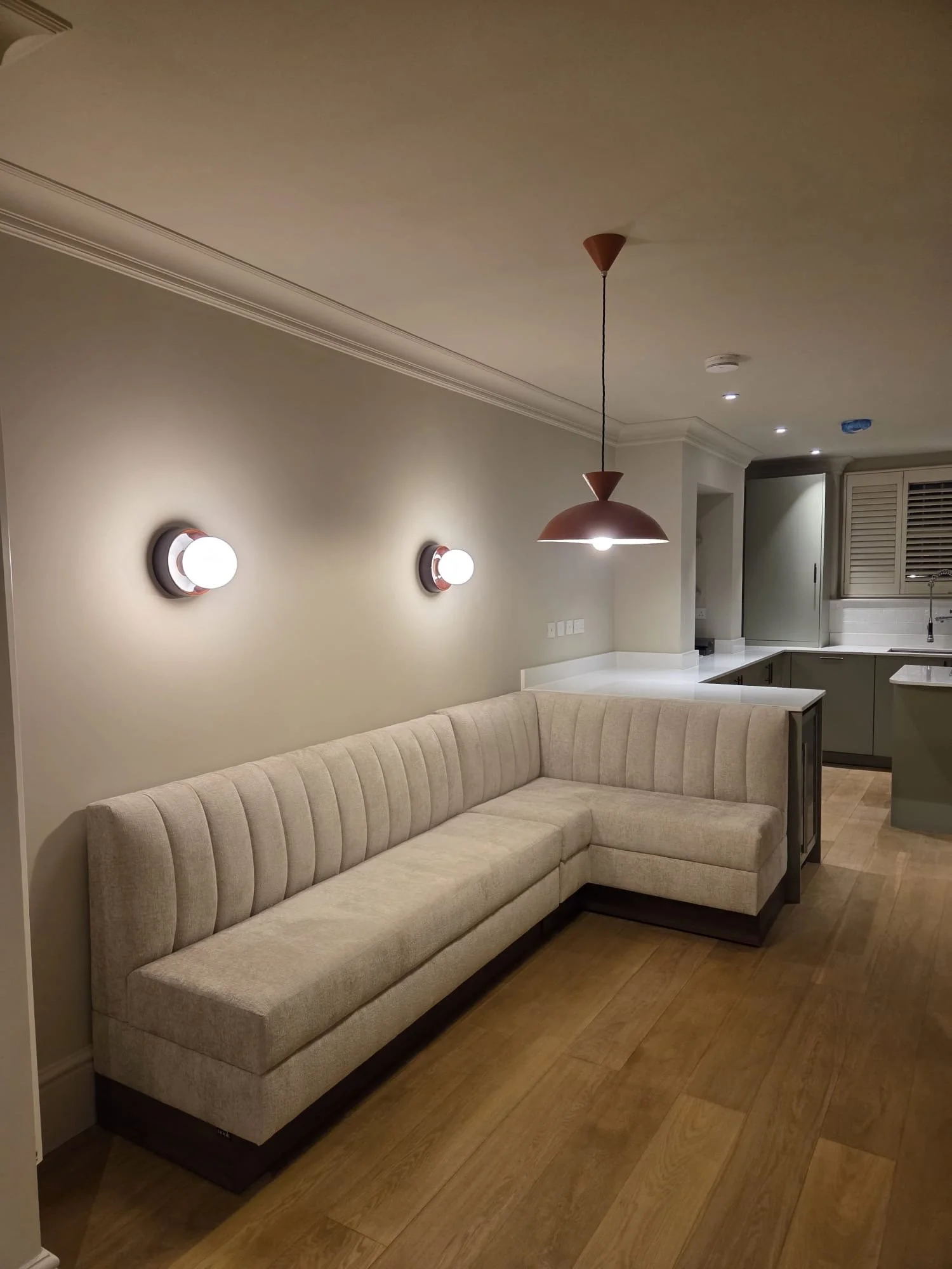 Living room with beige sectional sofa, two wall-mounted lights, and a hanging pendant lamp over the sofa in a modern kitchen.