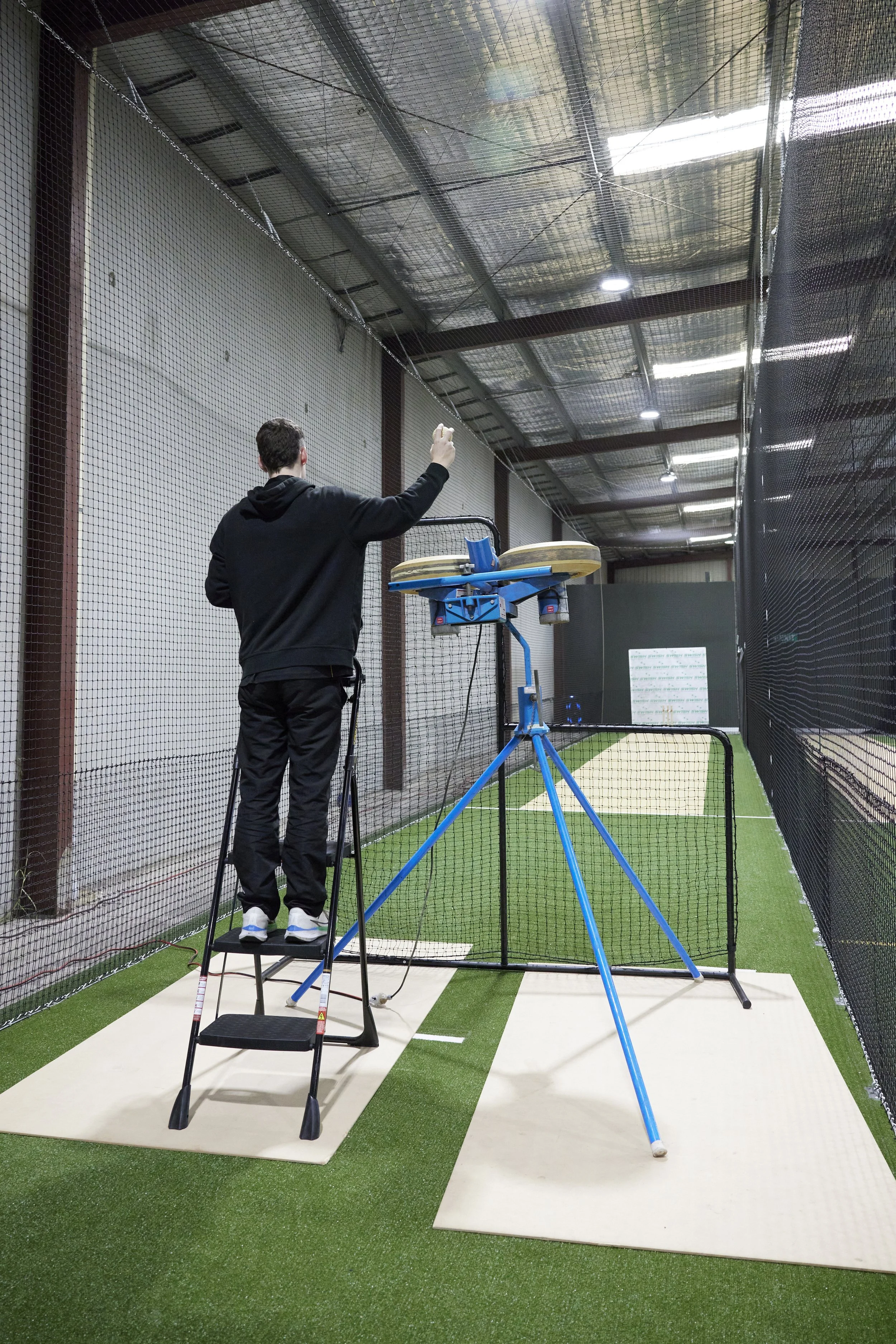 Person standing on a stool inside a sports training area, preparing to hit a cricket ball from a pitching machine.