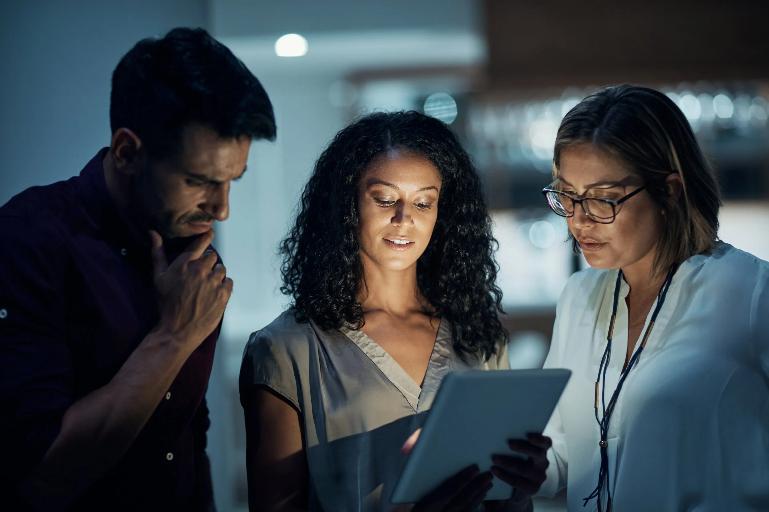 Three professionals reviewing content on a tablet together in a dimly lit workspace, representing early adoption of Workday Developer Agent