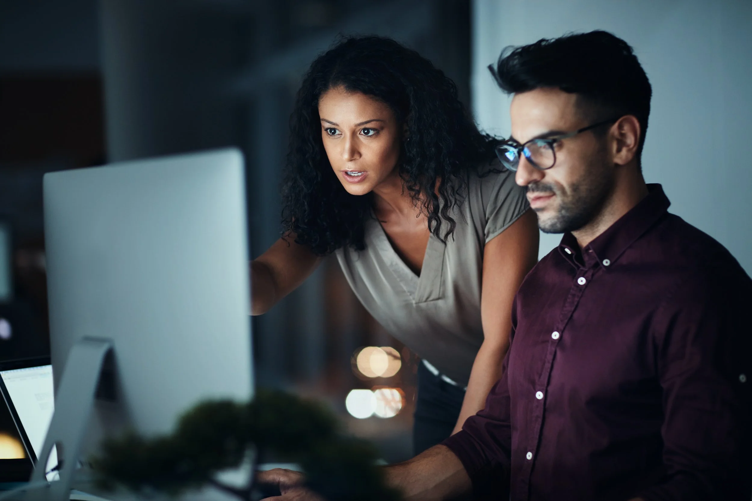 Two colleagues reviewing work together on a desktop monitor in a dimly lit office, representing Workday implementation consulting