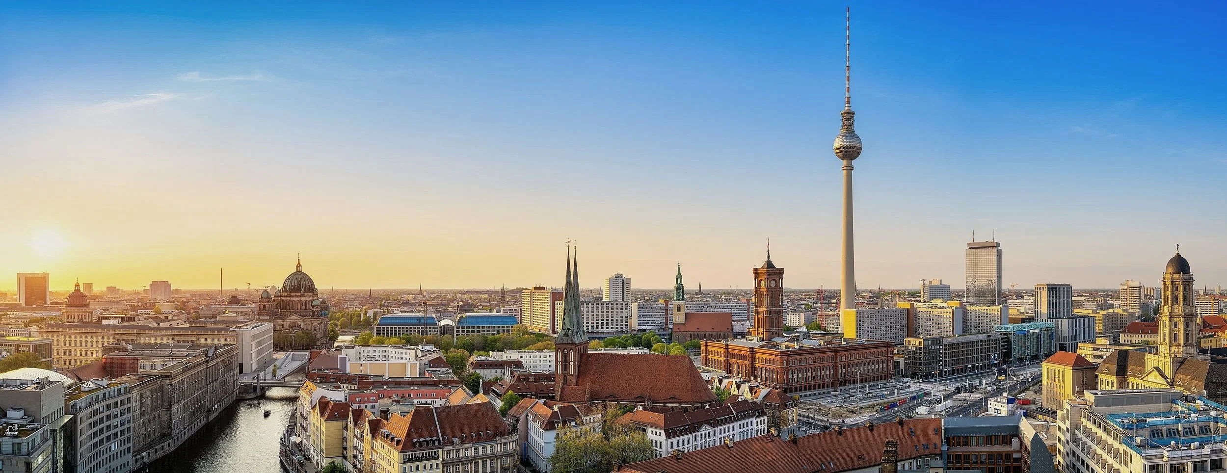 Berlin skyline at sunset with the Fernsehturm tower above city rooftops under a clear blue sky.