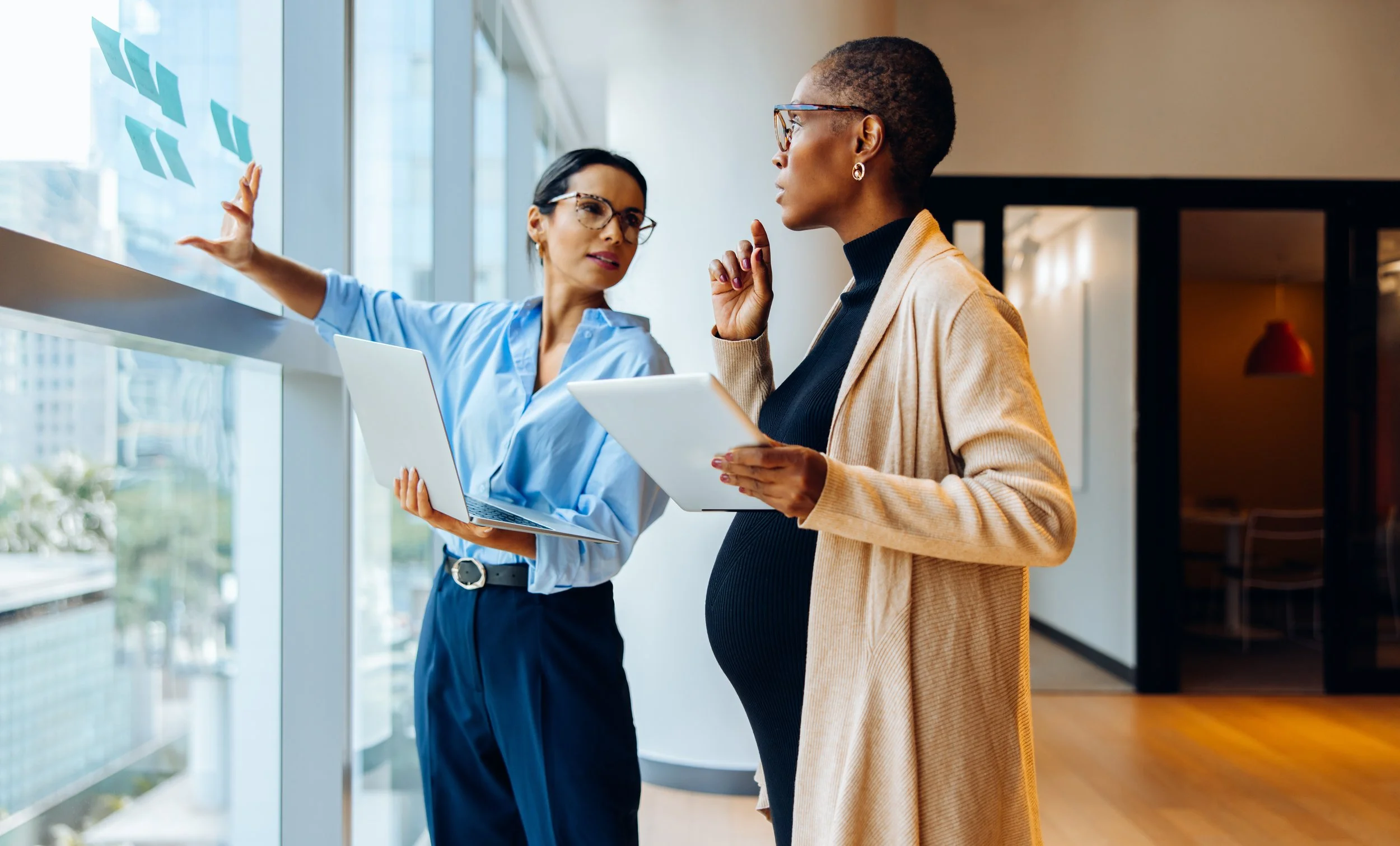 Two professionals reviewing sticky notes and discussing ideas beside a window in an office