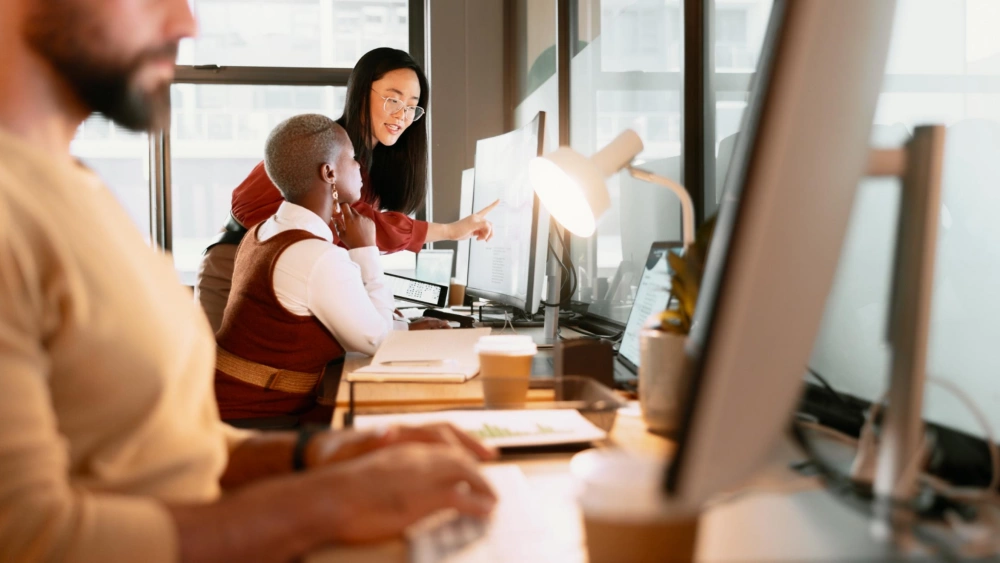 In a bright open-plan office, a colleague stands beside a seated coworker and points at a large computer monitor while they review something together; other desks and screens are visible in the foreground.