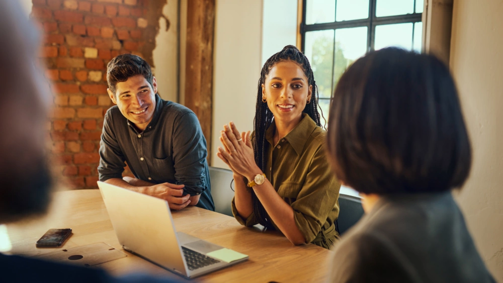 Three colleagues in a meeting room sit around a table with a laptop; one person speaks with hands clasped while another smiles, with a brick wall and window in the background.