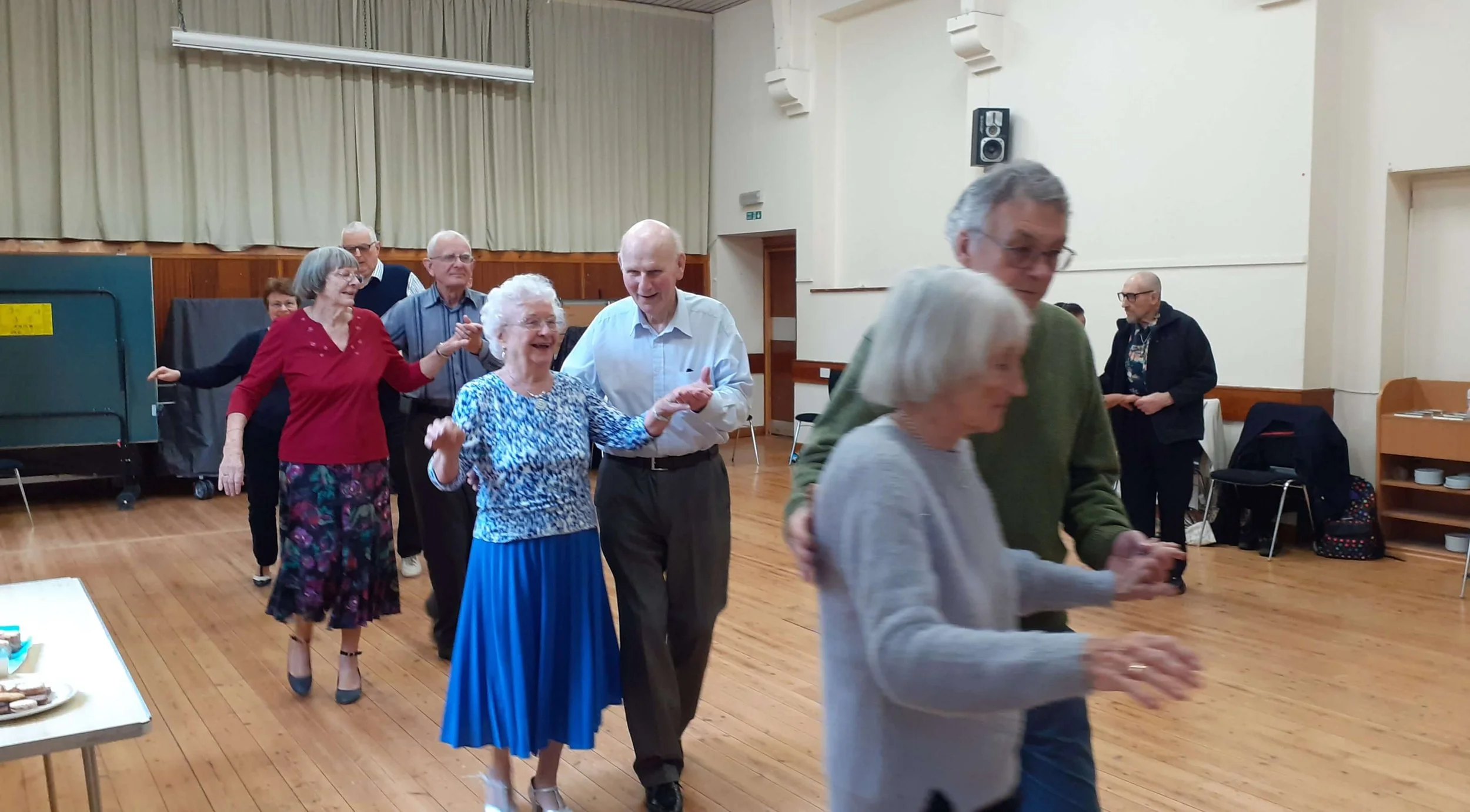 A village hall with curtains and high ceilings where older people are dancing in pairs