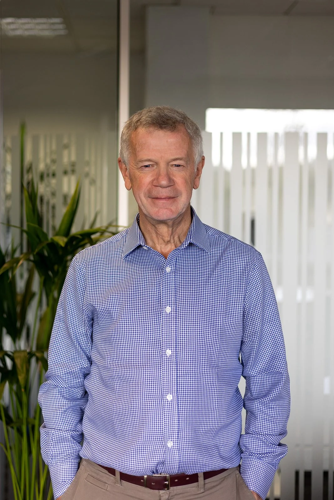 Nick stands smiling in a blue checked shirt with his hands in his pockets. He is a slim white man with grey hair.