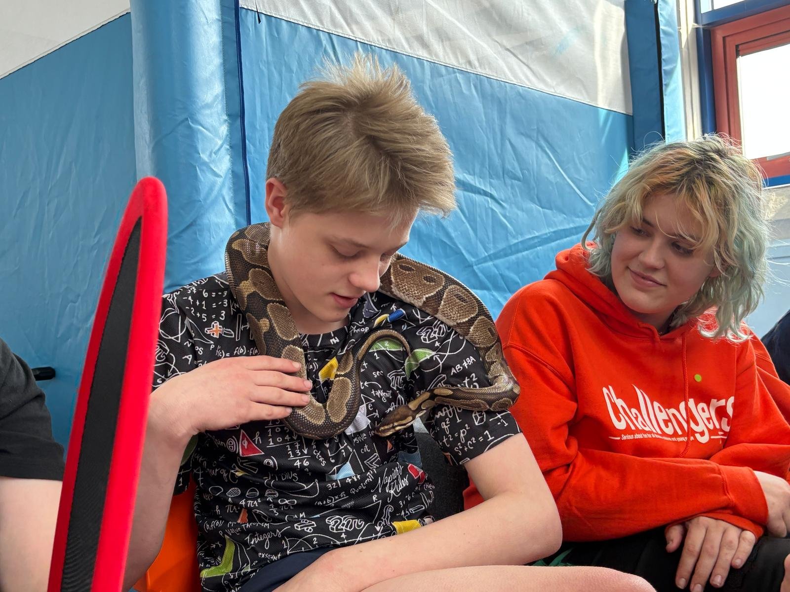 A boy sits with a Royal Python over his shoulders next to a woman with curly blonde hair in an orange Challengers hoody