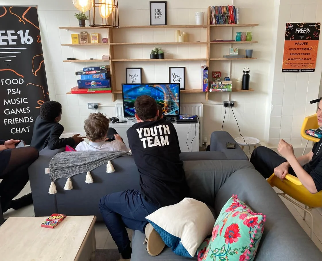 A group of young people pictured from behind sitting playing on a console in a white room with modern shelving. A man in a black tshirt with "YOUTH TEAM" written on the back leans on the back of the sofa to join in.
