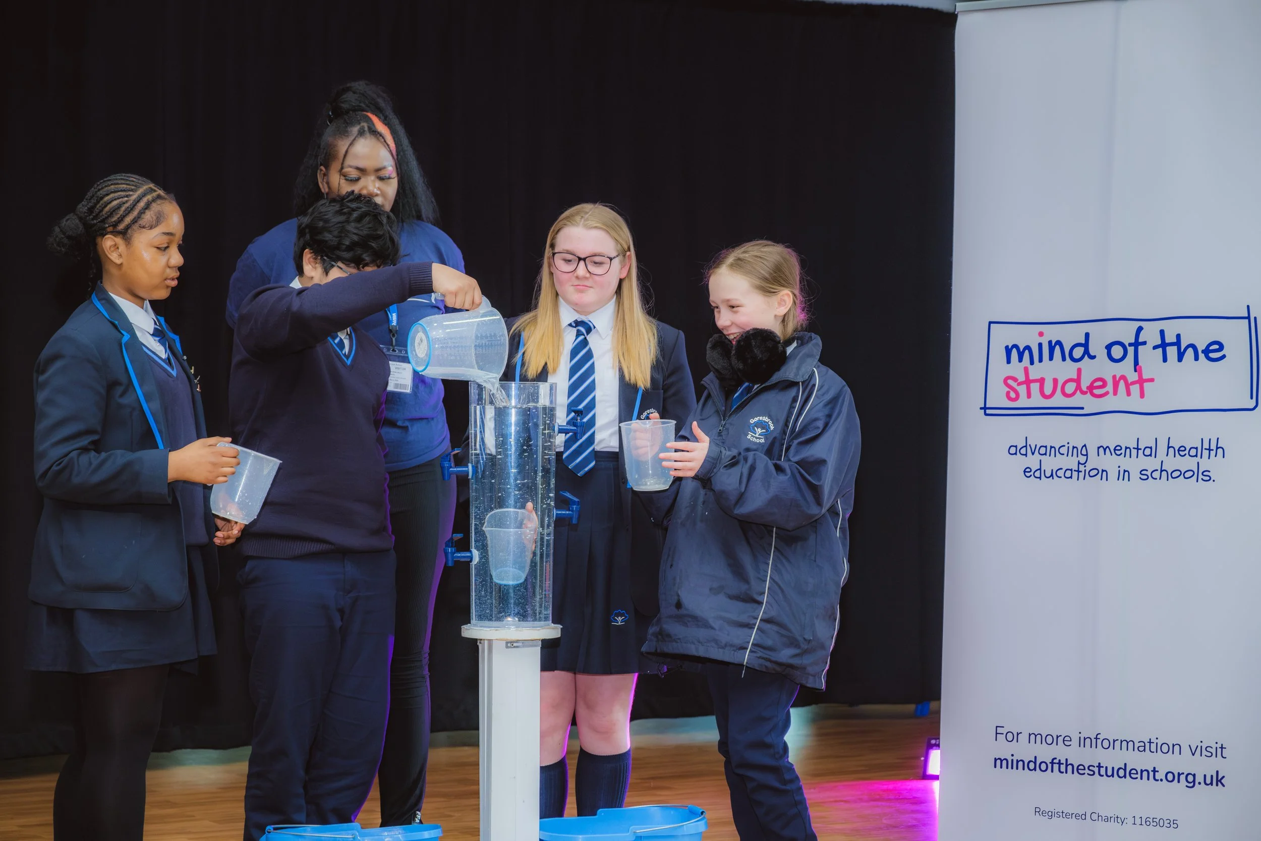 Students at a Surrey school are taking part in an exercise about mental health, pouring water into a cylinder with a jug each. There is a Mind of the Student banner beside them.