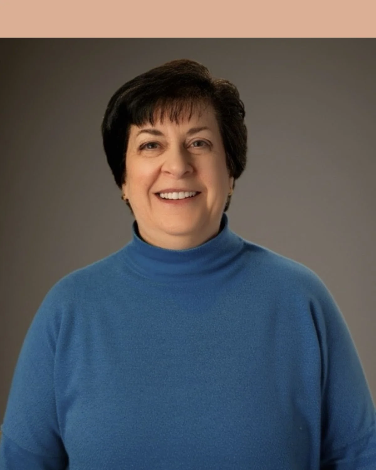 A woman with short dark hair, wearing a blue turtleneck sweater, smiling and standing against a tan brick wall.