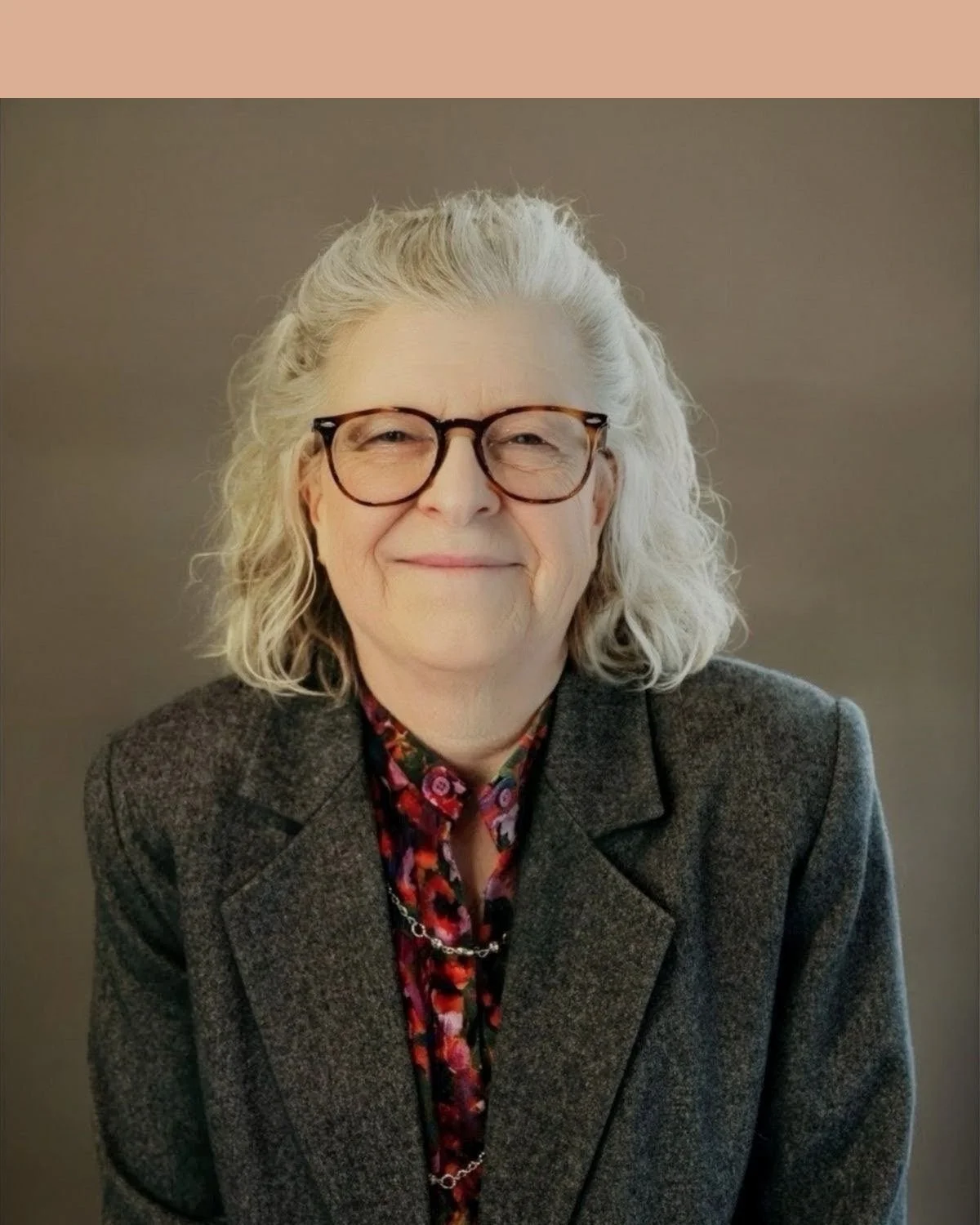 Smiling older woman with curly gray hair wearing glasses, a dark floral shirt, and a gray blazer standing against a beige brick wall.