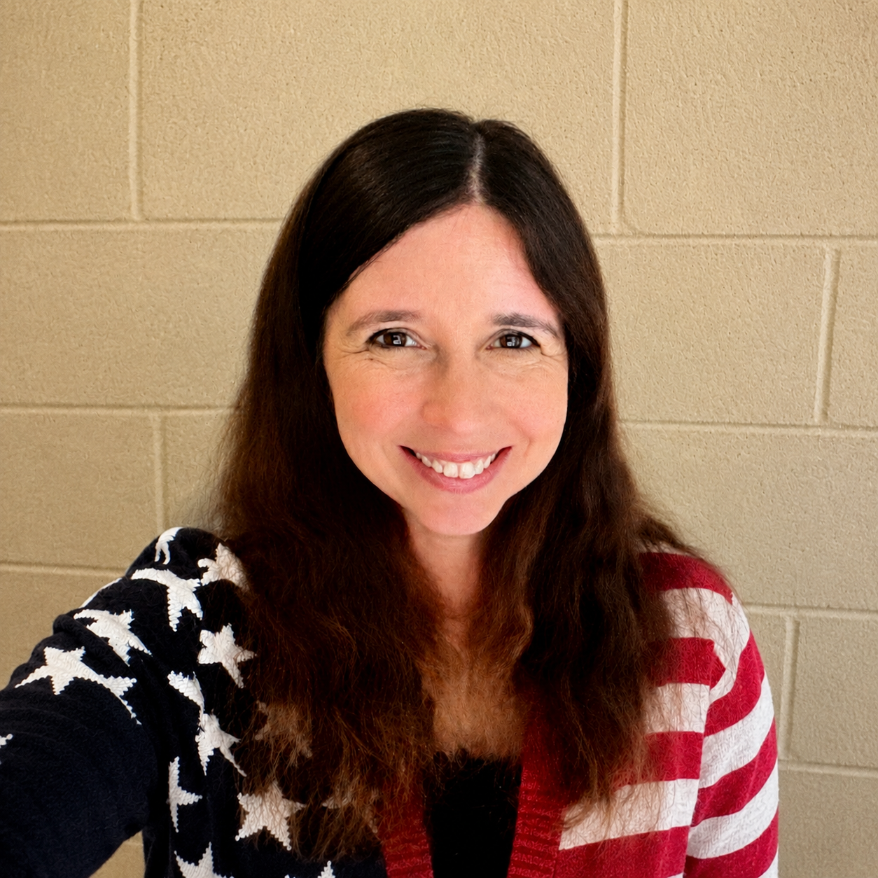 A woman with long dark brown hair smiling at the camera, wearing a jacket with American flag pattern.