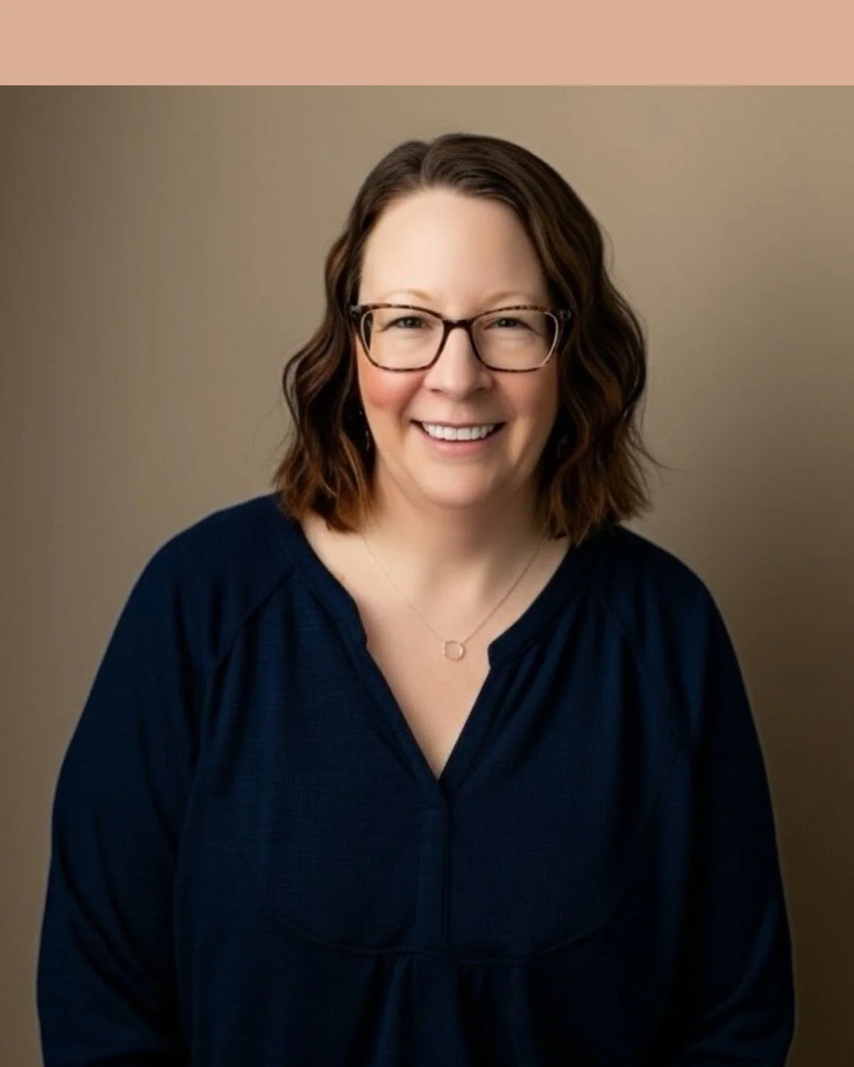A woman with shoulder-length brown wavy hair, wearing glasses and a navy blue textured blouse, smiling in front of a beige cinder block wall.