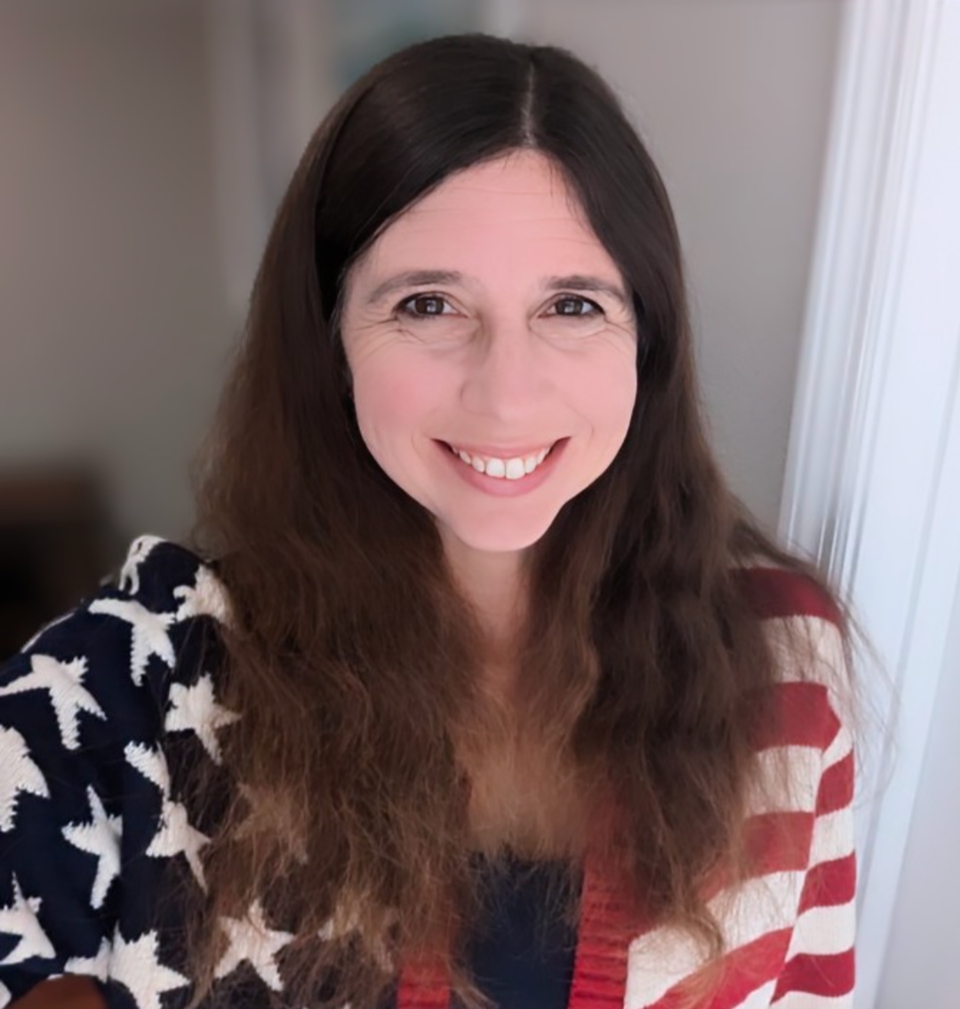 A woman with long dark brown hair smiling at the camera, wearing a jacket with American flag pattern.