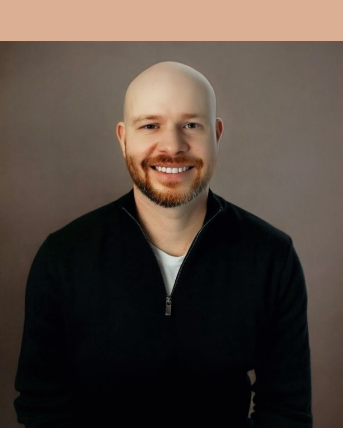 A smiling man with a beard and a bald head, wearing a black zip-up sweater over a white shirt, sitting against a beige brick wall.
