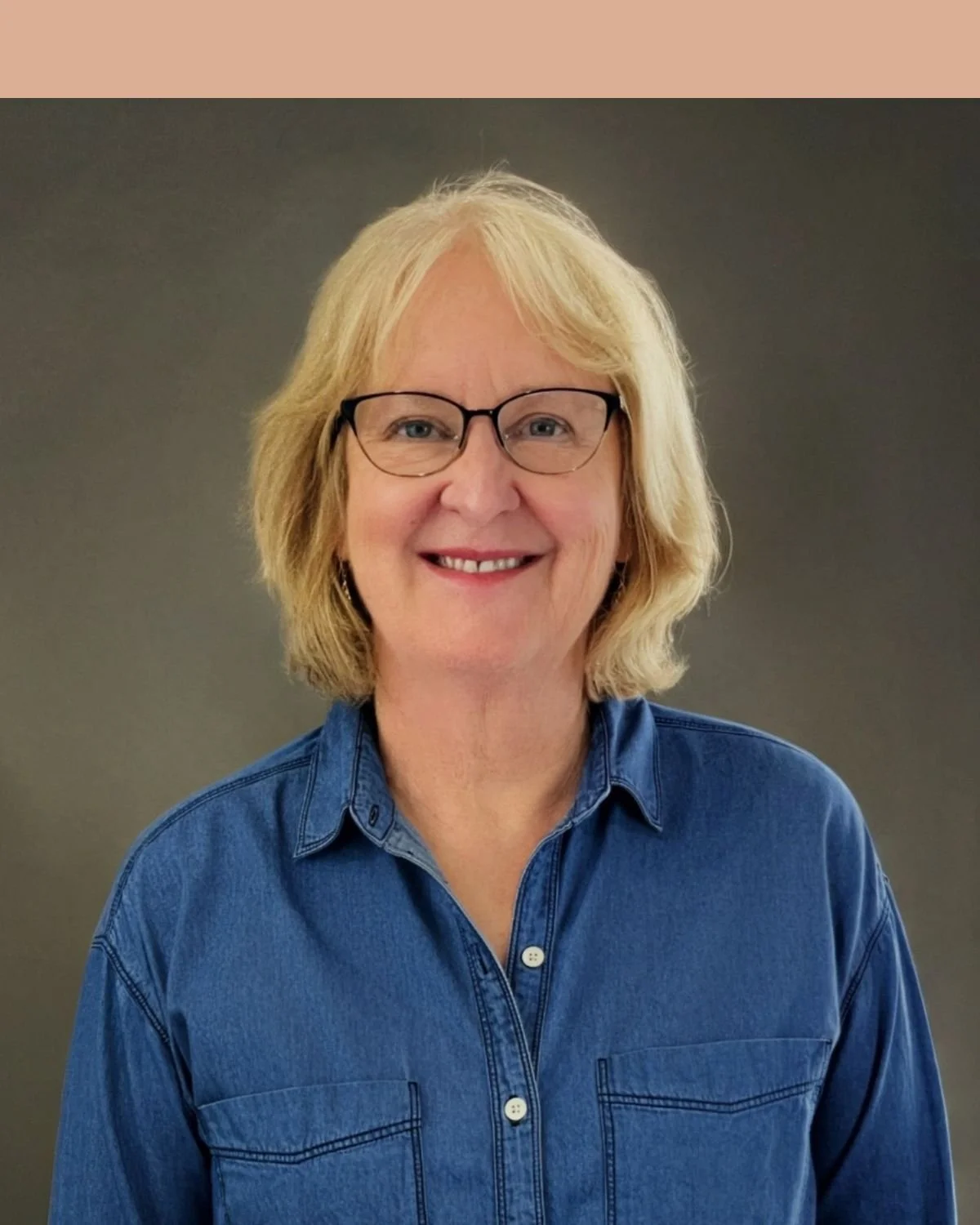 A smiling woman with blonde hair and glasses, wearing a textured royal blue top, standing against a beige brick wall.