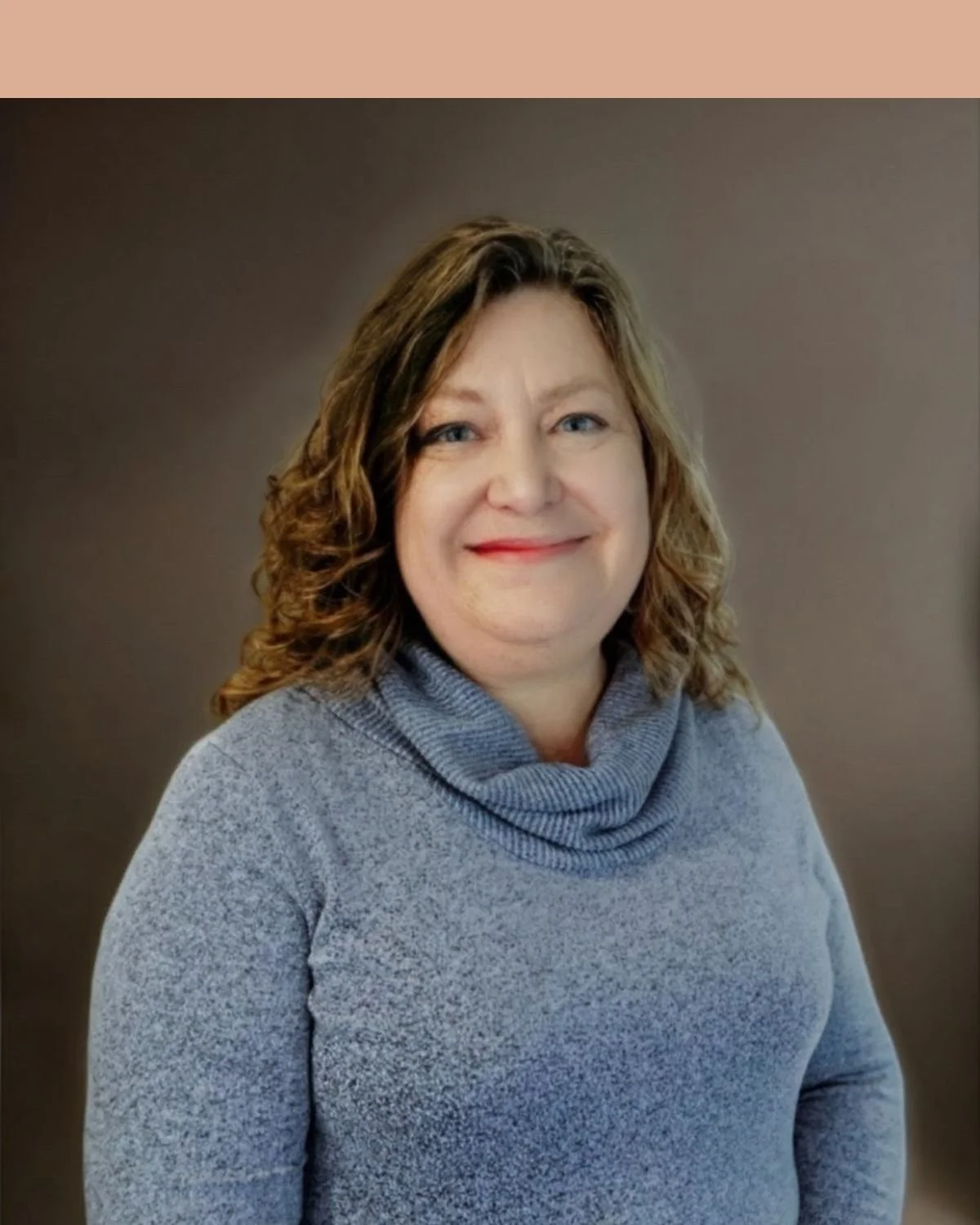 A woman with shoulder-length, wavy brown hair and a light complexion, smiling, standing against a beige brick wall, wearing a gray turtleneck sweater.