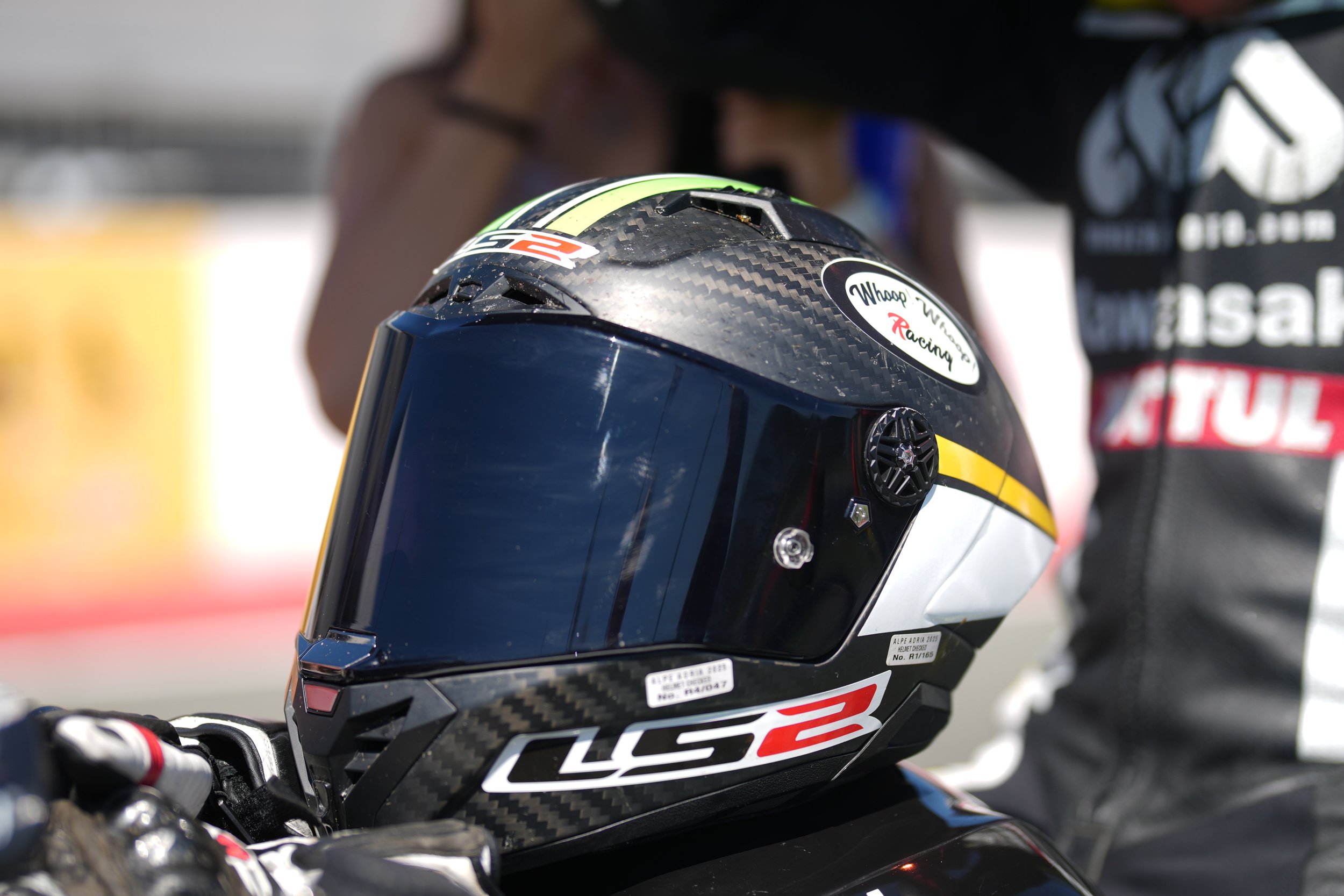 Close-up of a black racing helmet with the label "LCS2" and various stickers, positioned on a motorcycle handlebar, with a blurred background of a person and racing environment.