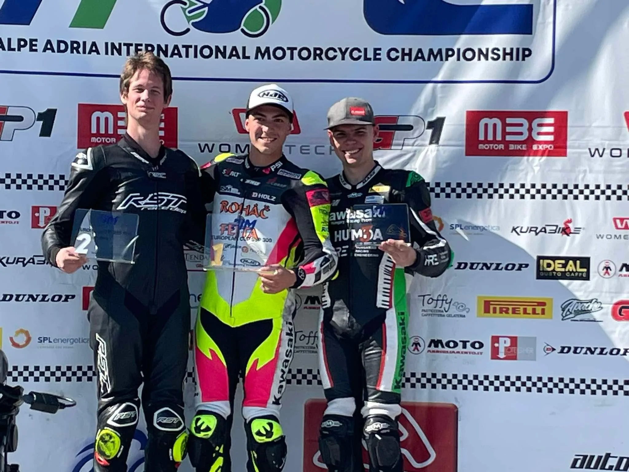 Three motorcycle racers on a podium at the Alps Adriatic International Motorcycle Championship, holding trophies, with a backdrop displaying logos and sponsors.