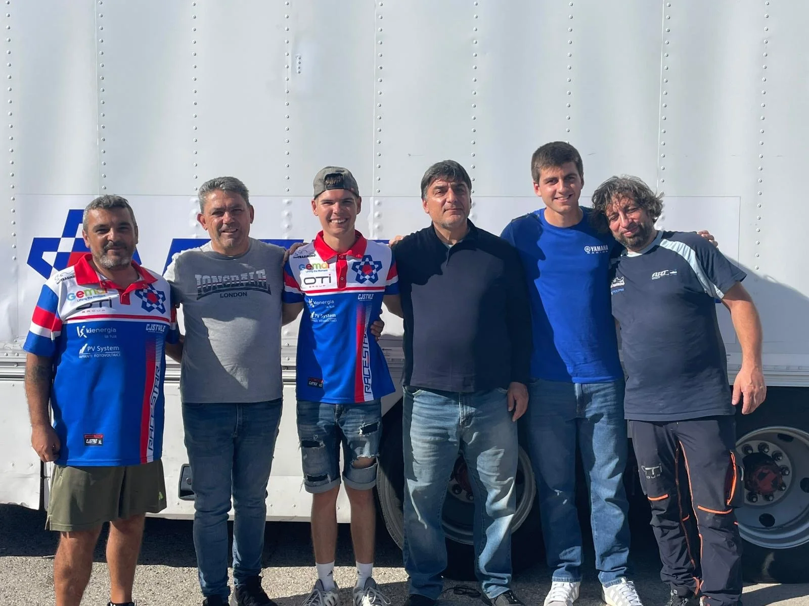 Group of six men standing outdoors, smiling, with a large white bus behind them. Three of the men are wearing matching blue and white racing jerseys.