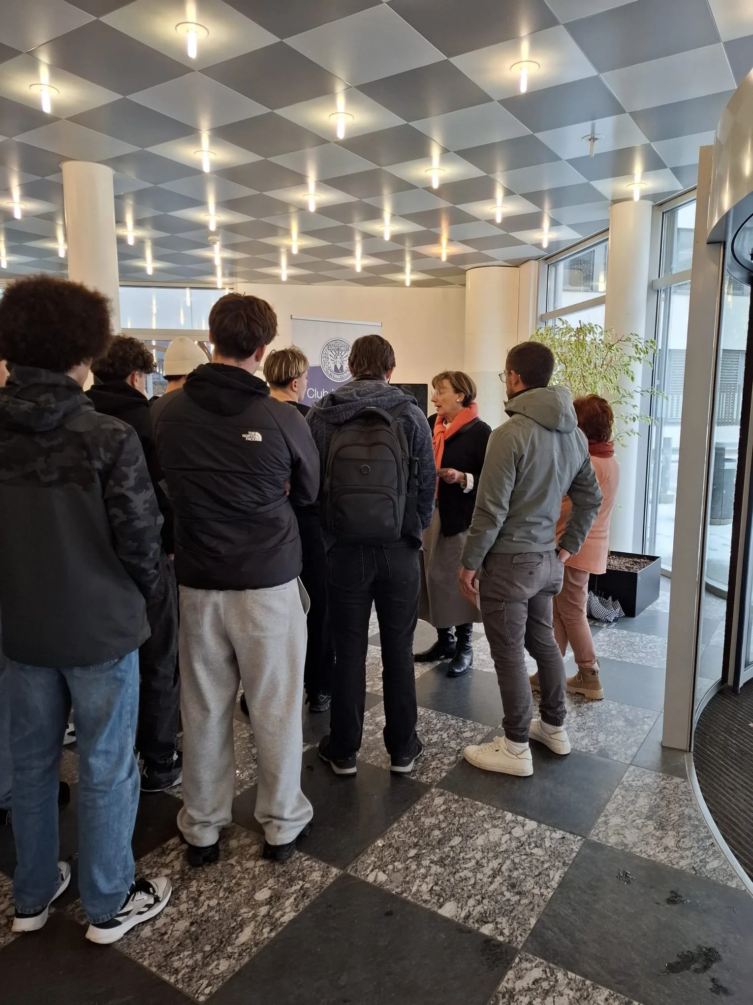 Groupe de personnes attendant ou discutant dans un endroit intérieur avec un plafond suspendu et de grandes fenêtres. Une femme parle à un groupe, une autre personne porte un sac à dos.