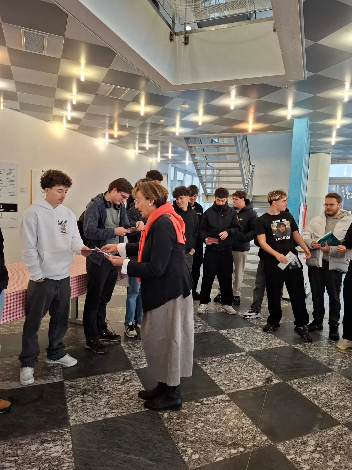 Groupe de jeunes hommes et femmes en file d'attente dans un hall moderne, discutant avec une femme qui leur remet des documents ou des brochures.