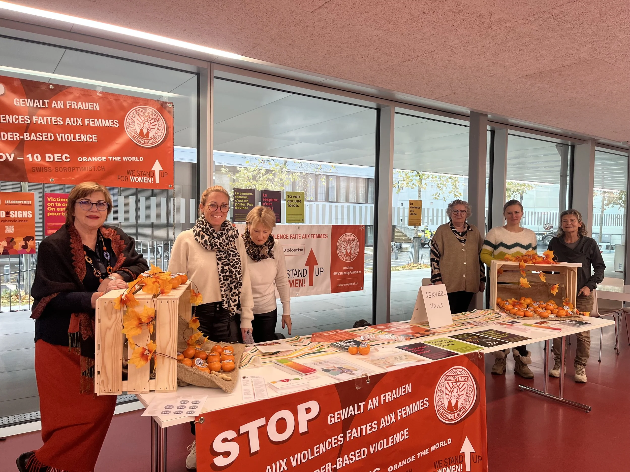 Groupe de six femmes derrière une table d'information lors d'une manifestation contre la violence à l'égard des femmes, avec des affiches en plusieurs langues et une bannière en arrière-plan en français et en anglais.