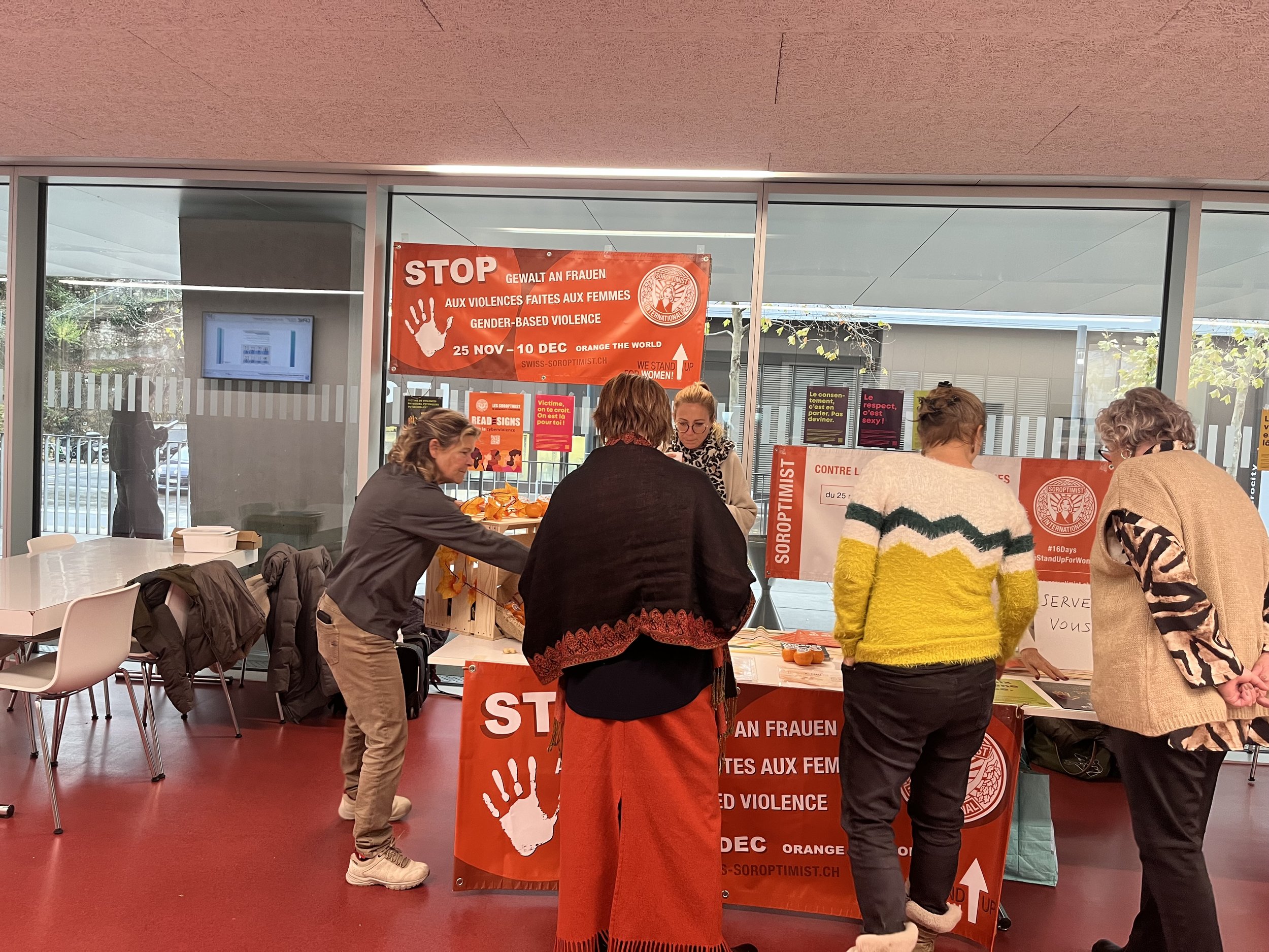 Groupe de personnes à une table d'information en lutte contre la violence faite aux femmes, dans un espace intérieur avec affiches en français et en anglais.