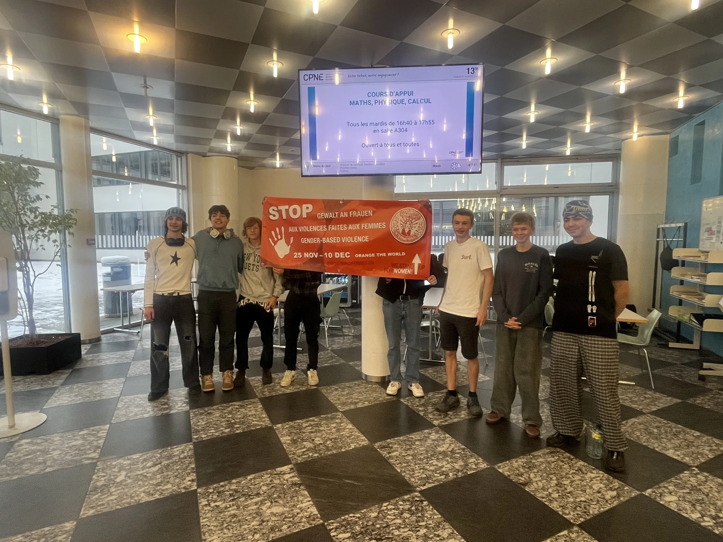 Groupe de sept jeunes debout dans un hall, tenant une bannière rouge pour la sensibilisation contre la violence basée sur le genre, avec une montre digitale suspendue au plafond au-dessus d'eux.