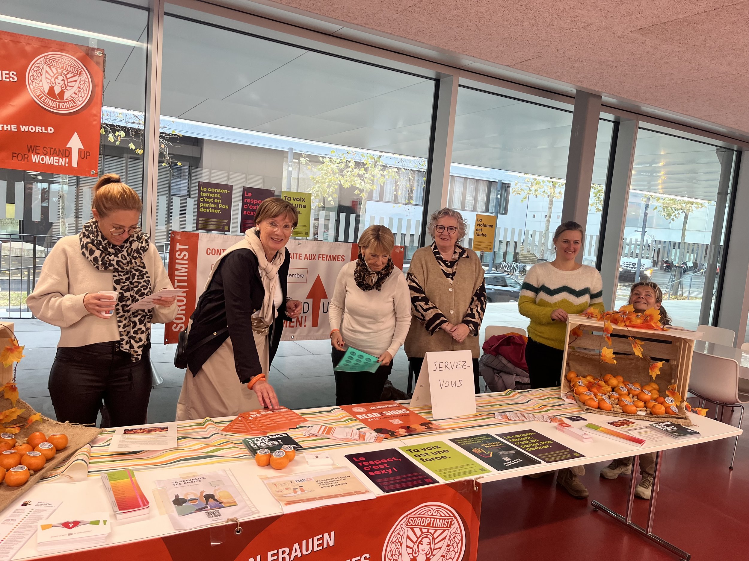 Groupe de femmes à un stand de sensibilisation sur la violence faite aux femmes, avec affiches, brochures, et boîtes de mandarines