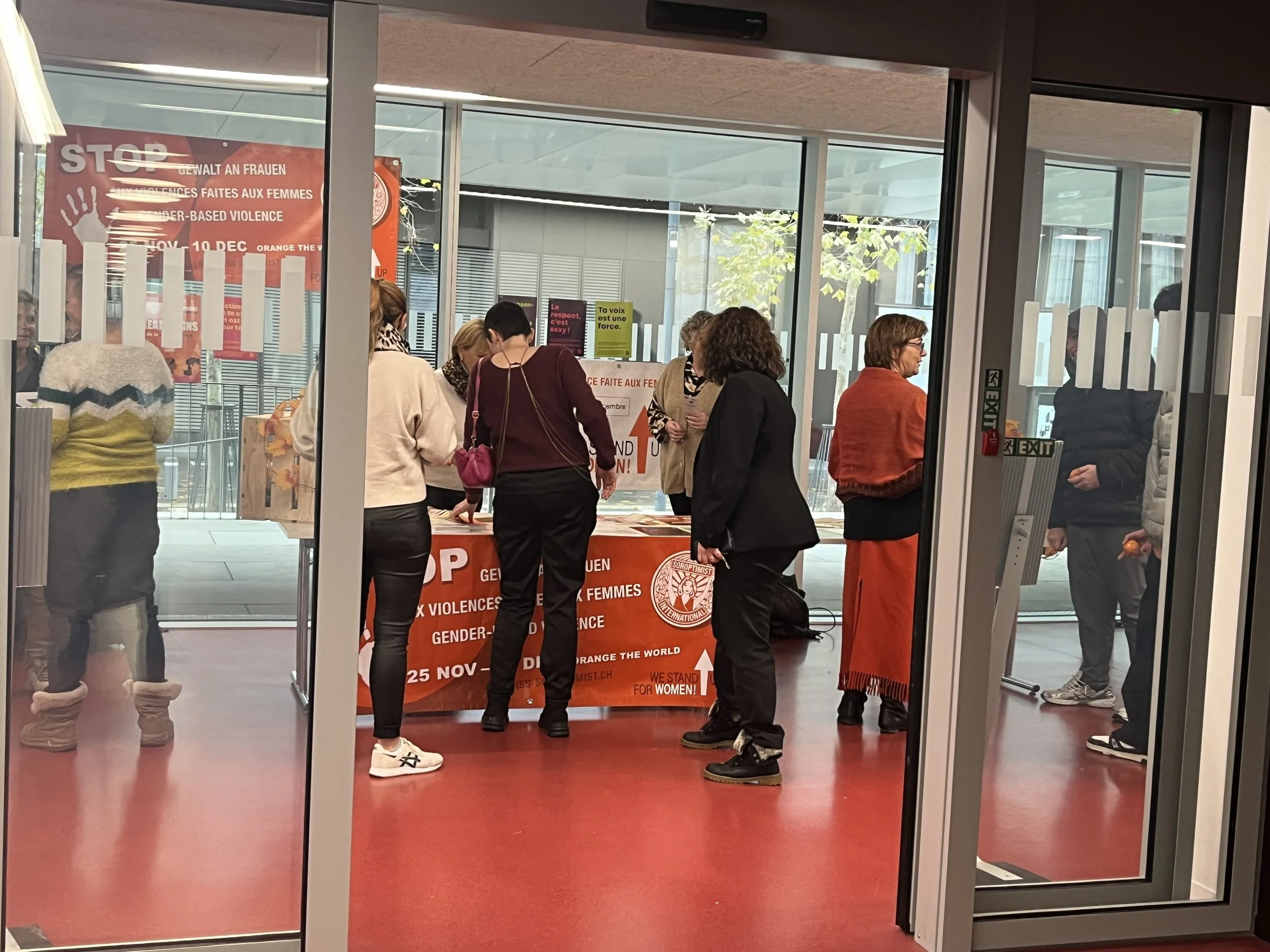 Groupe de personnes dans une salle, participant à une manifestation ou un événement contre les violences faites aux femmes. Un panneau orange en arrière-plan mentionne la date 25 novembre et le slogan "Stop aux violences faites aux femmes".