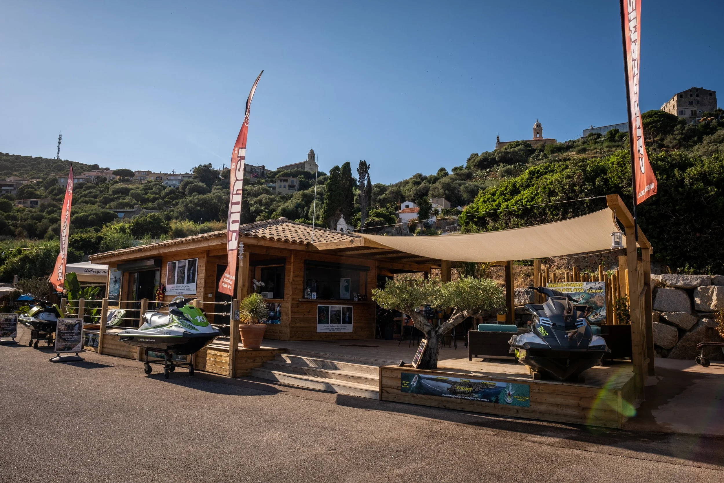 Station de jet ski en bois avec drapeaux orange, situé en zone touristique avec collines verdoyantes en arrière-plan, sous un ciel clair et ensoleillé.