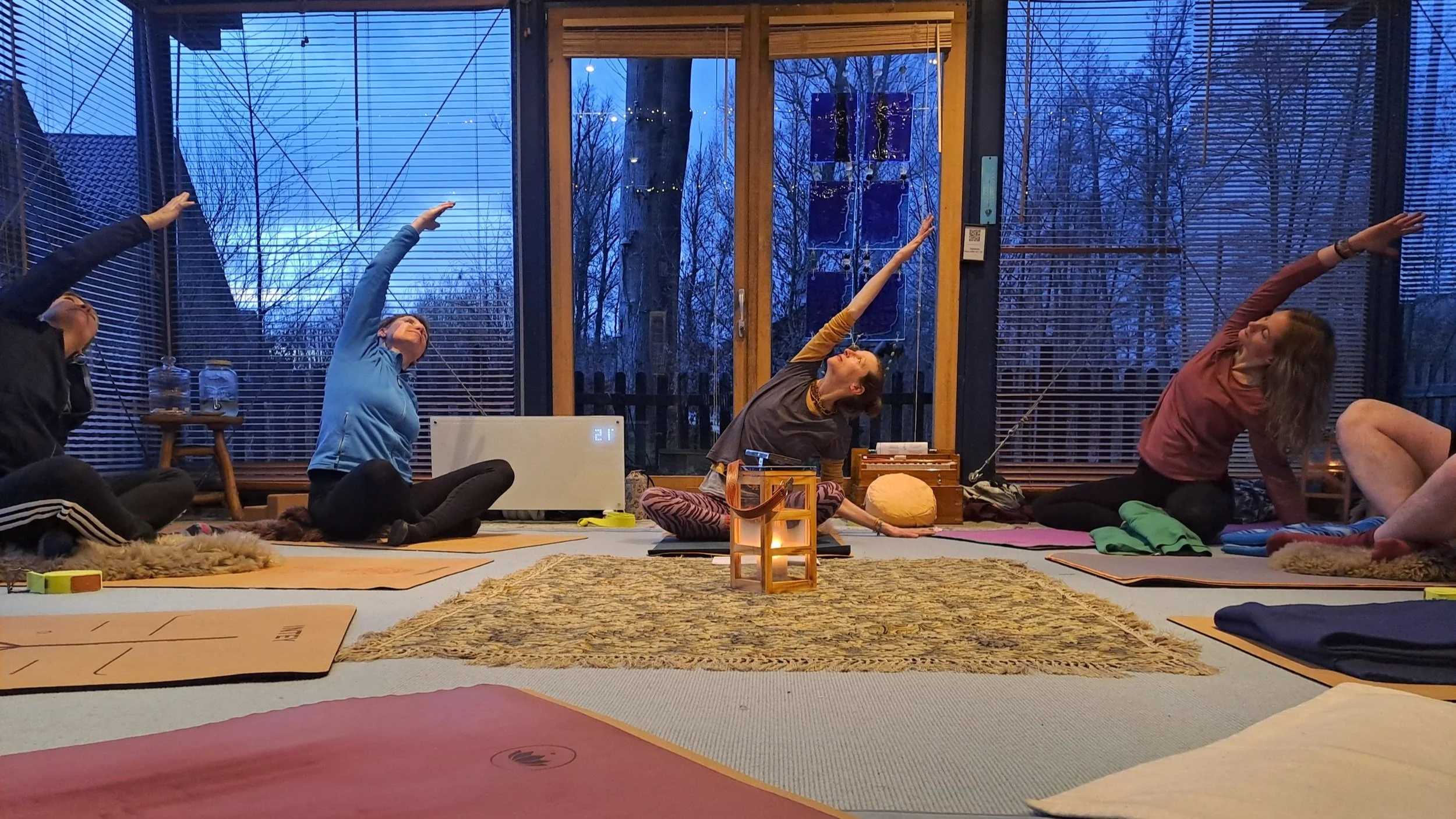 4 Frauen sind auf Yogamatten in einem Raum mit großen Fenstern in einer Seitbeuge im gekreuzten Sitz (Aufwärmübung)