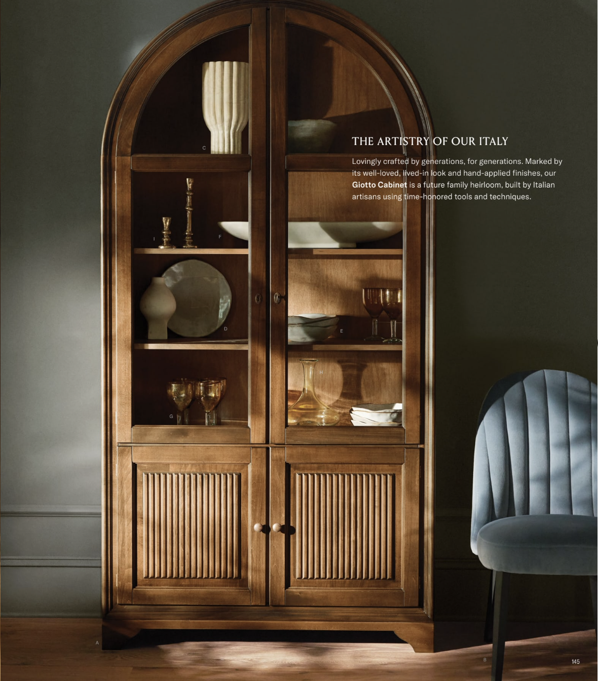 Arhuas 2024 Fall Catalog -- photo shows an arched wooden hutch with fluted details. inside are plates, candles, glasses, vases and more. a small periwinkle side chair sits next to it.