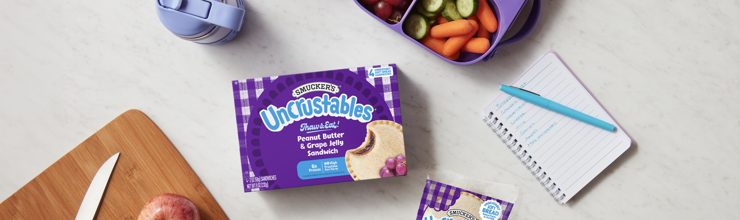 Uncrustables Campaign 2024 -- photo shows an uncrustables box with a cutting board, notepad and other snacks.