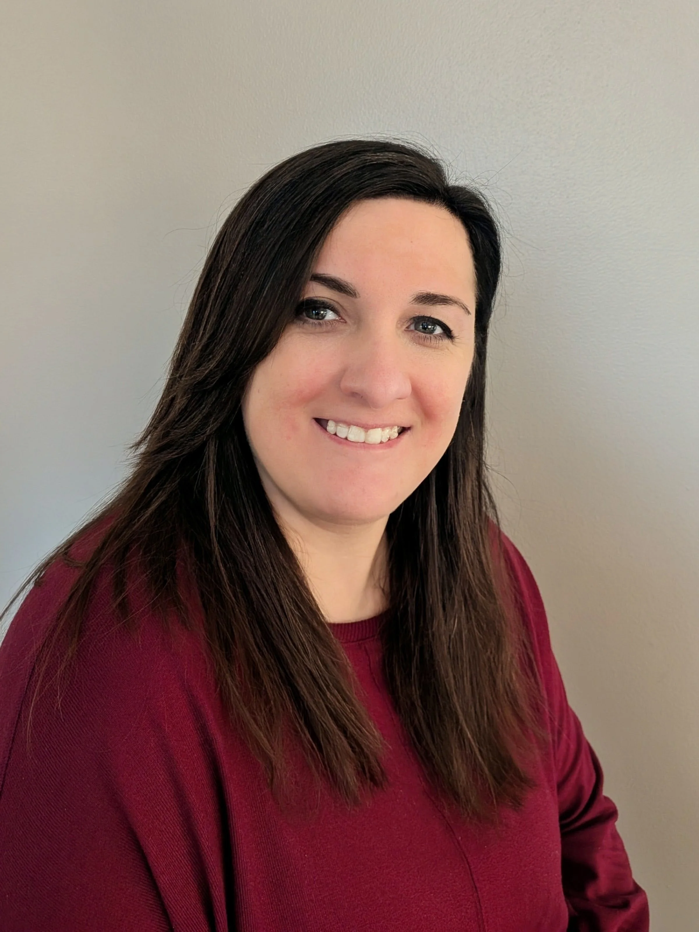 A woman with long dark hair smiling, wearing a maroon top, standing against a light-colored wall.