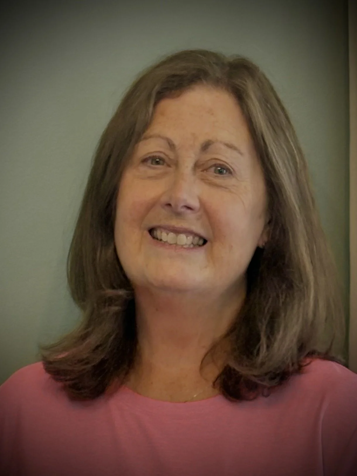 A woman with shoulder-length brown hair, smiling and wearing a pink top, poses in front of a plain background.