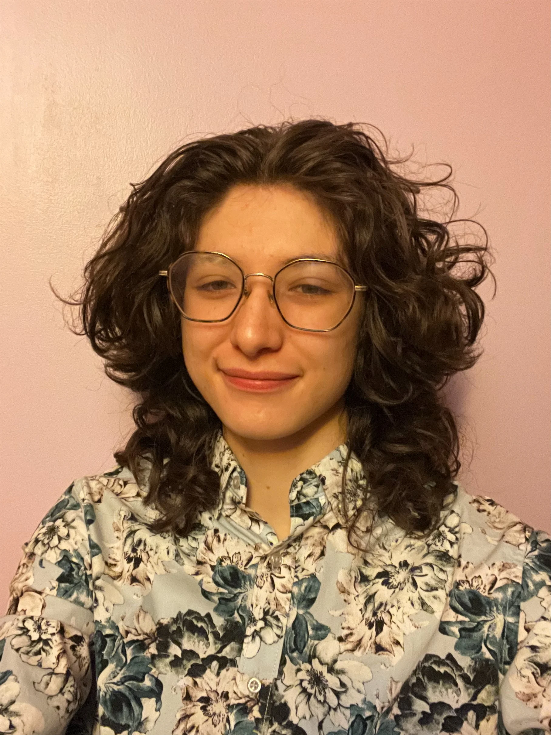 A young woman with medium skin tone, wavy dark hair, wearing glasses, a floral shirt, and a necklace, smiling in front of a pink wall.