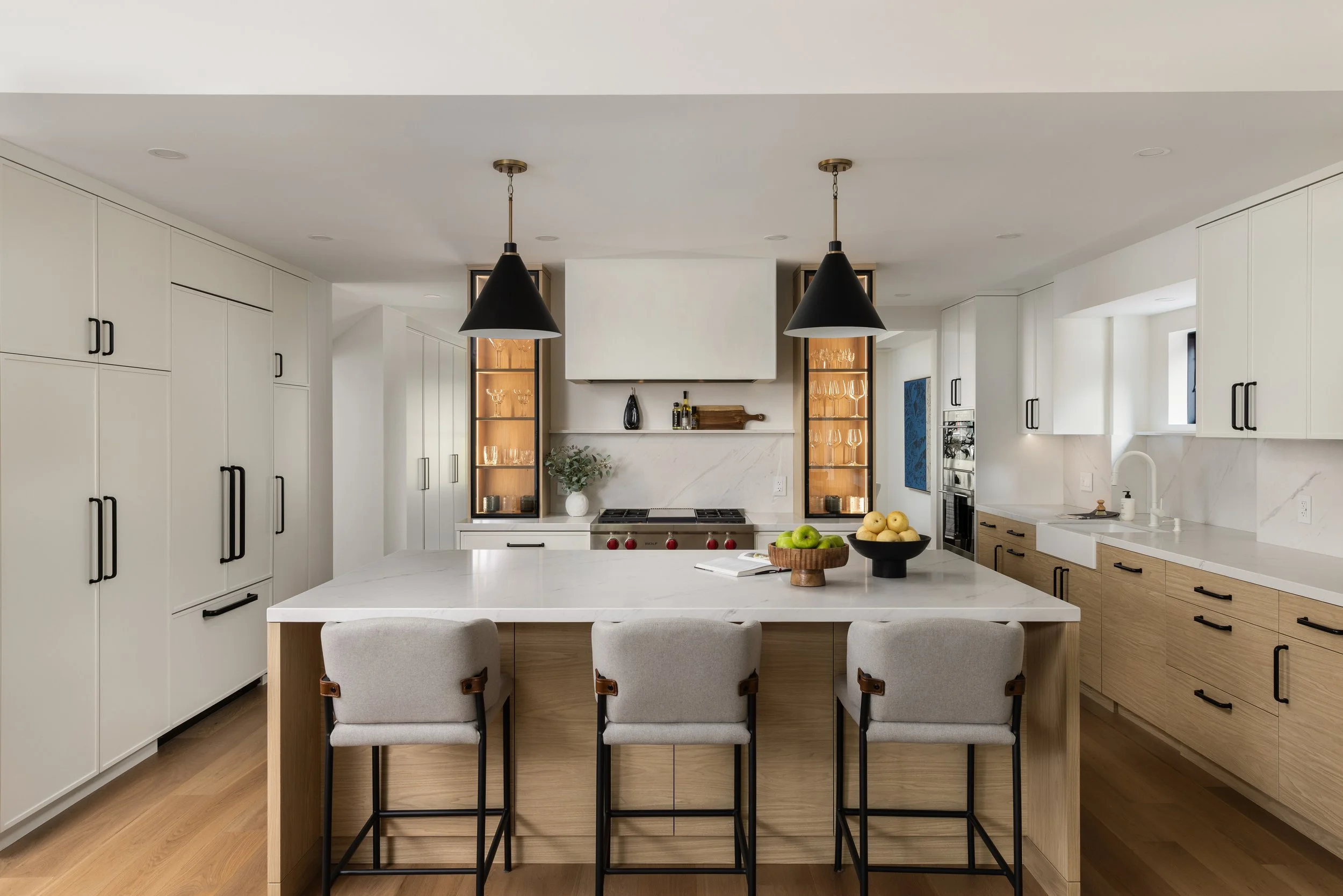 View of a bright modern kitchen from the open-concept living room, featuring a marble-topped kitchen island with three tall chairs on one side. A modern range is bordered by custom glass cabinets on either side and two conical black pendant lights